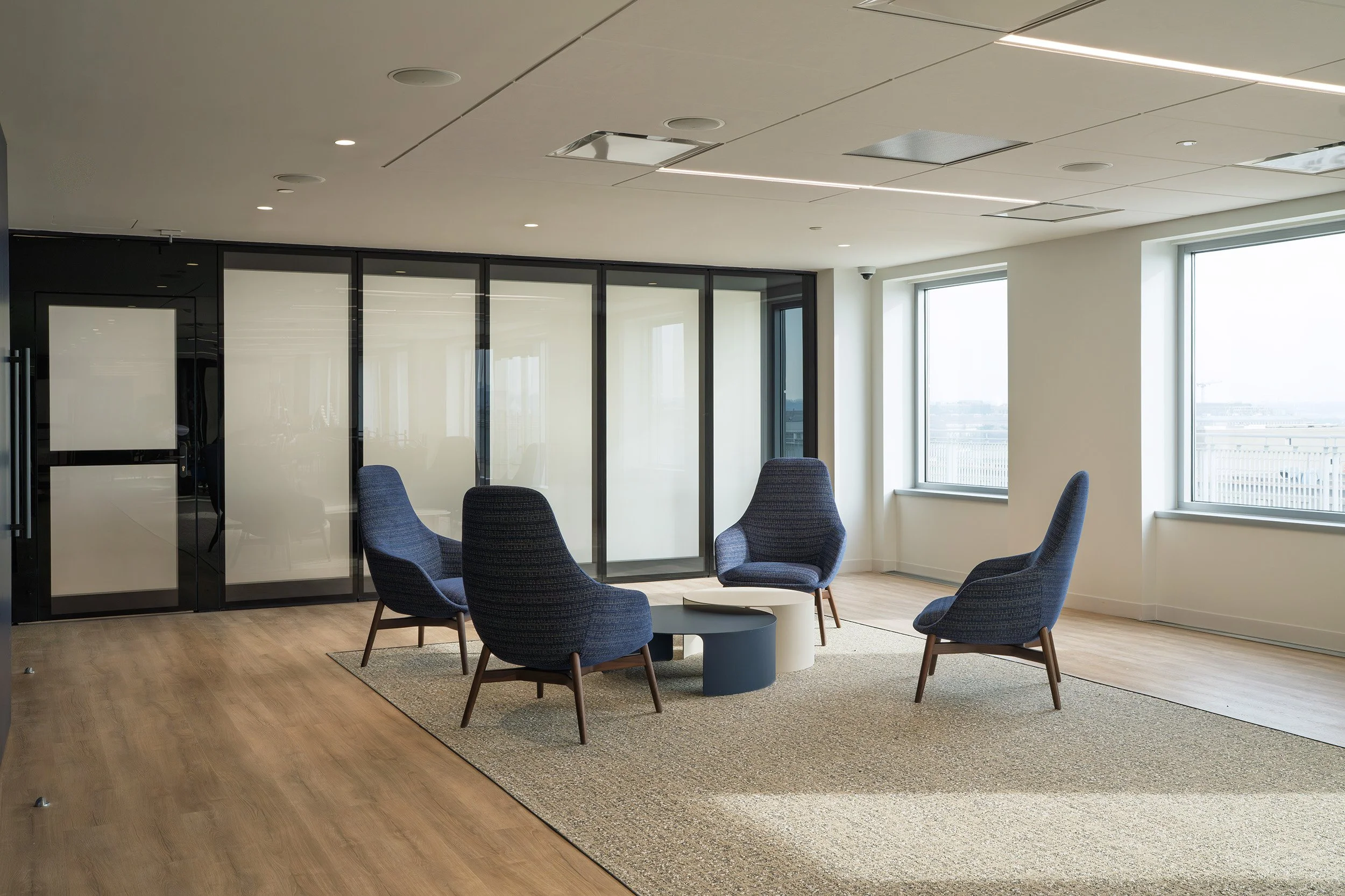 Modern office meeting area with four blue armchairs arranged around a round table on a beige rug, large windows, and glass partition walls.