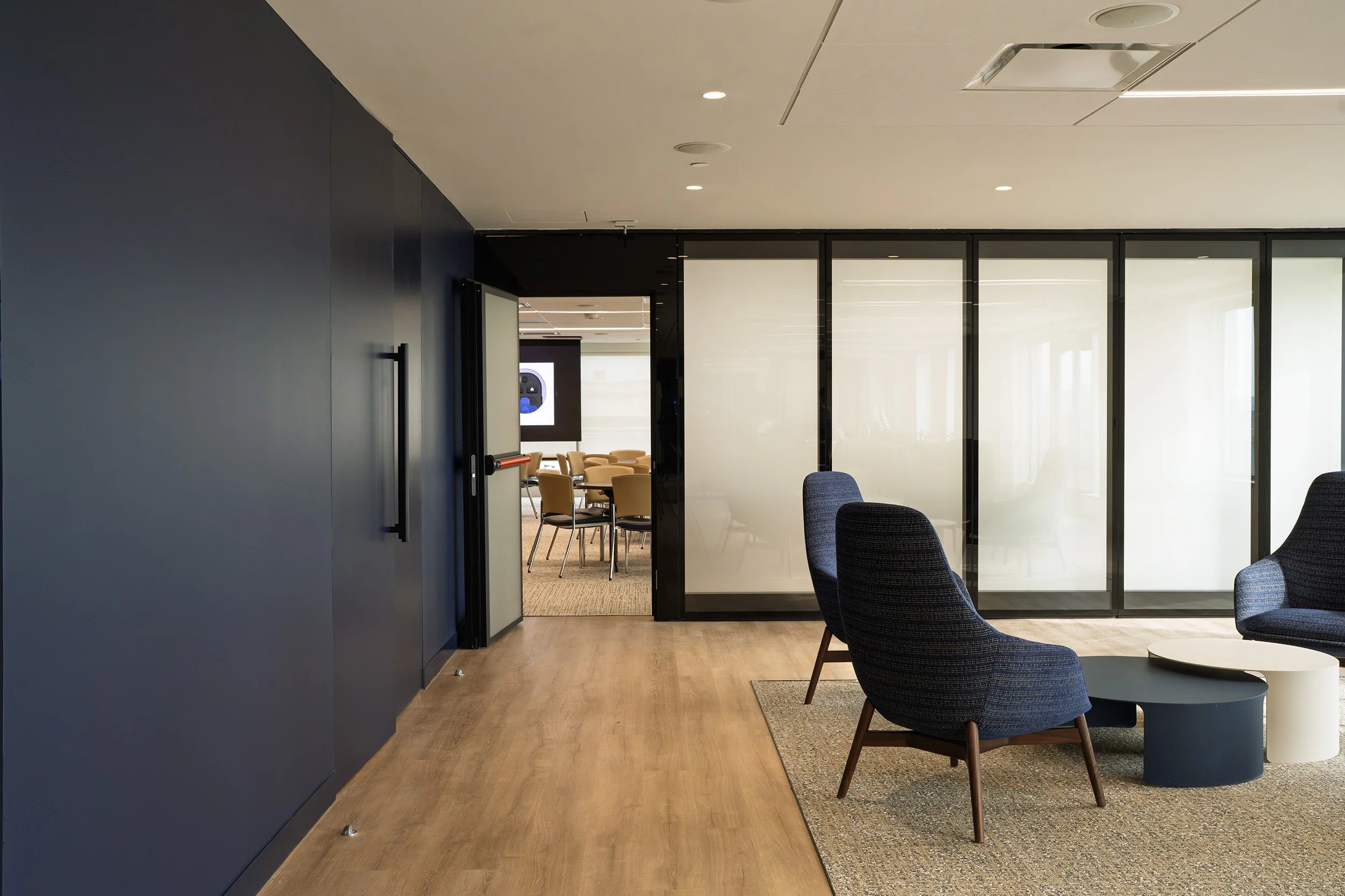 Modern office lounge with navy blue walls, wooden flooring, black-framed glass partitions, dark gray chairs, and a small round coffee table on a rug.