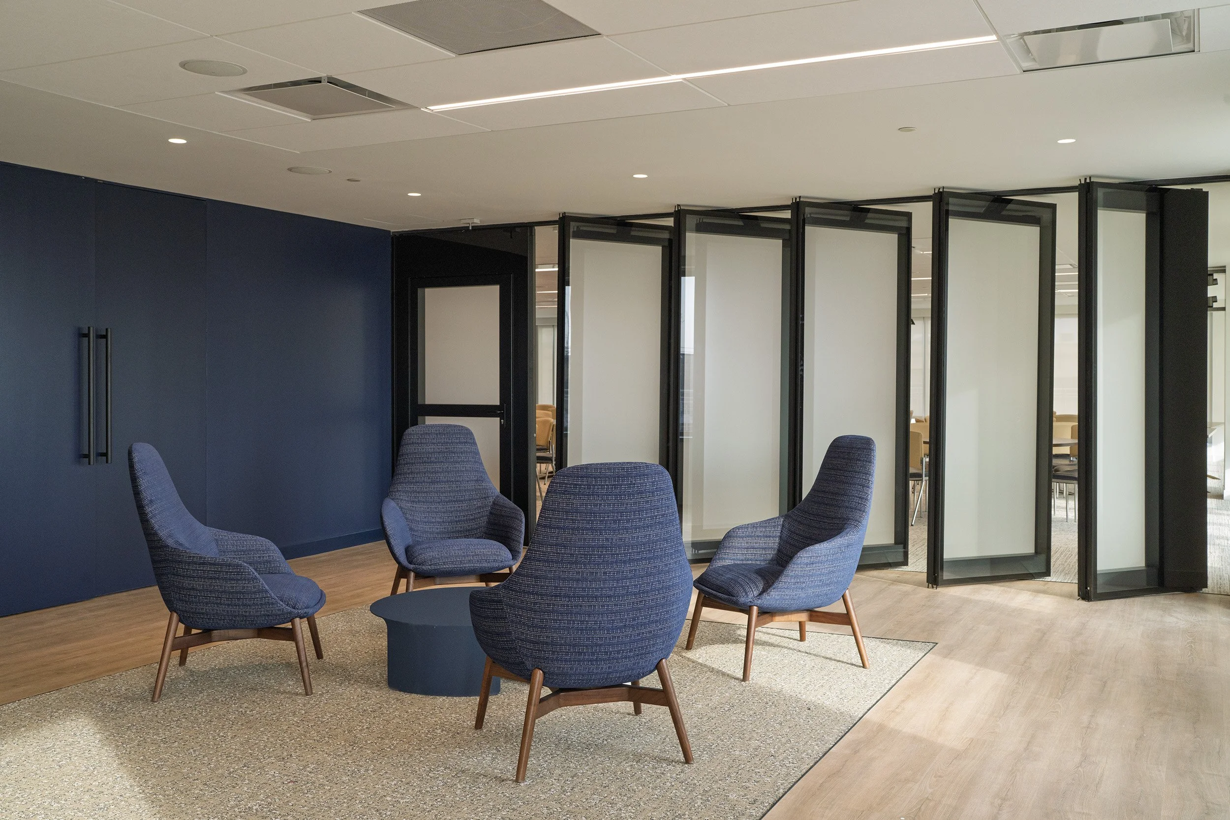 A modern office lounge area with four blue upholstered chairs around a small round table on a beige rug. In the background, there are folding glass panel partitions and a navy blue wall with black handles on a cabinet.