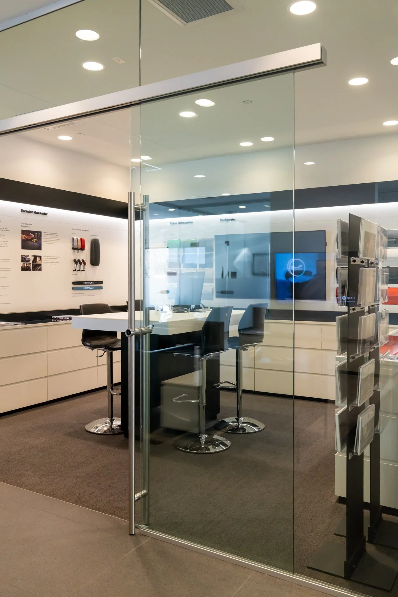 Office meeting room with a glass door, bar stools, a white table, black chairs, multiple monitors, and a display on the wall.