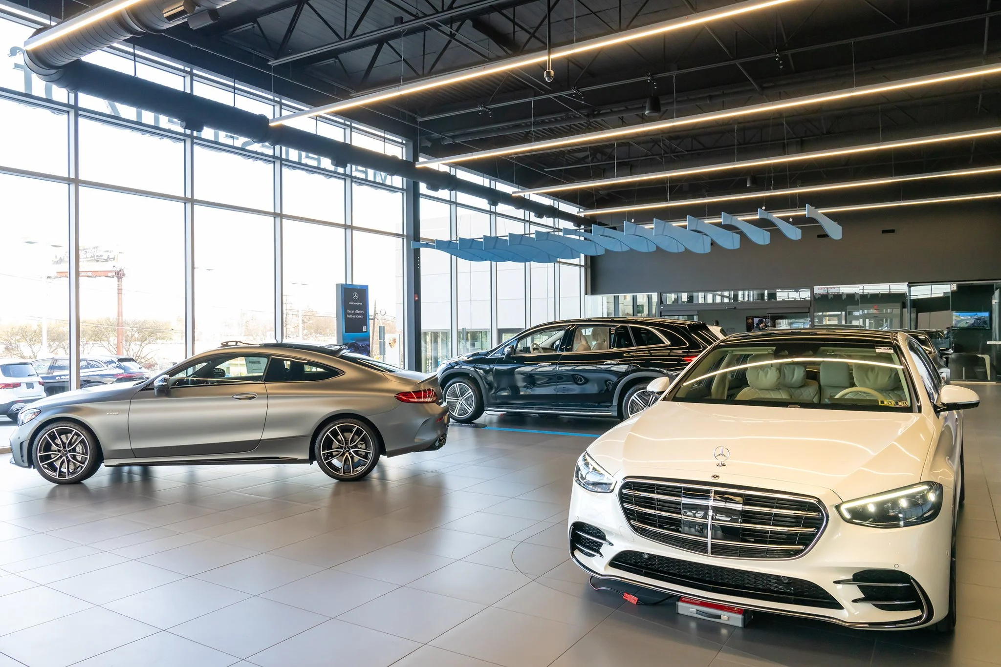 Interior of a luxury car dealership showroom featuring three Mercedes-Benz vehicles: a silver coupe, a black SUV, and a white sedan with a prominent front grille. Large windows let in natural light, and modern lighting fixtures are installed on the c