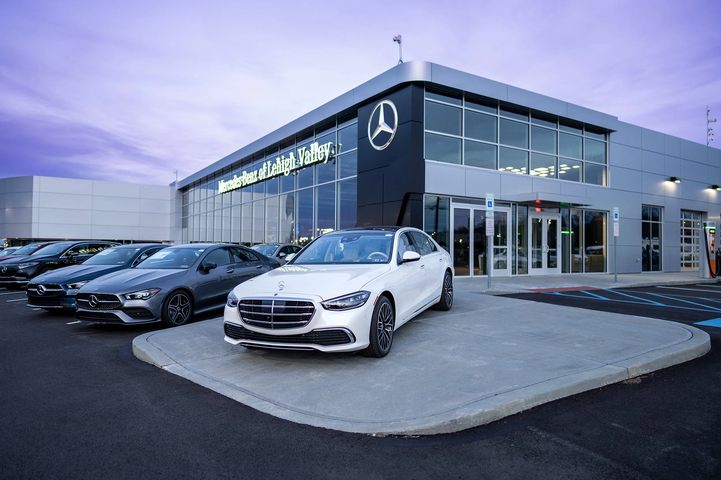 Exterior view of Mercedes-Benz dealership with several cars parked outside, including a white car prominently in front, and signage indicating Mercedes-Benz of Lehigh Valley during twilight.