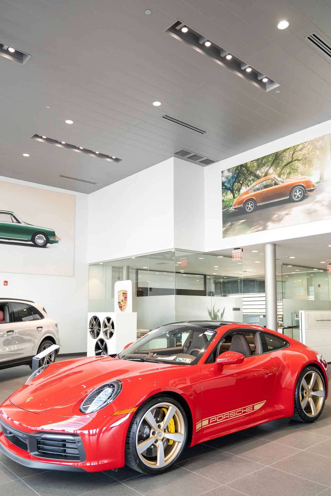 Red Porsche sports car on display in a showroom, surrounded by other cars and large automotive photographs on the walls.