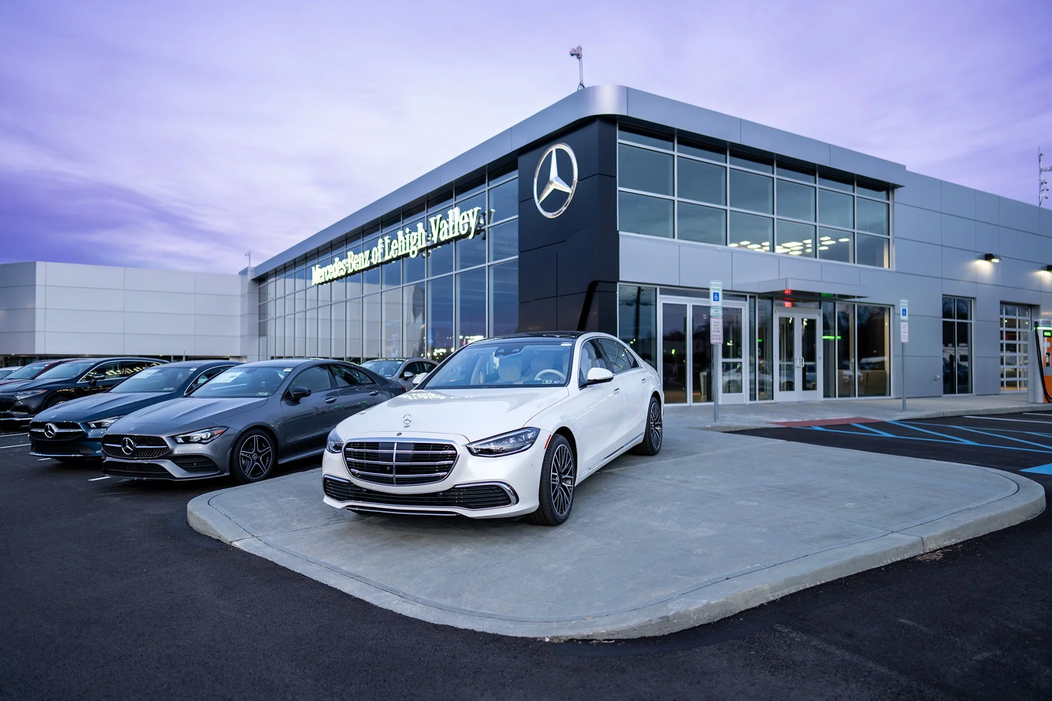 Mercedes-Benz dealership with multiple cars parked outside, including a white Mercedes on the sidewalk, and a modern, glass-front building with a Mercedes logo, under a purple evening sky.
