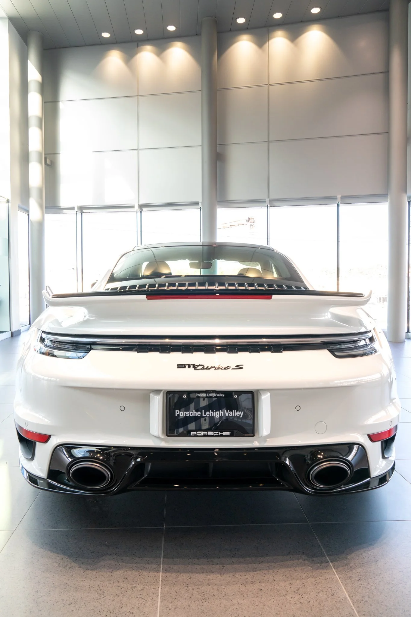 Front view of a white Porsche 911 Turbo S inside a modern showroom with large windows and ceiling lights.