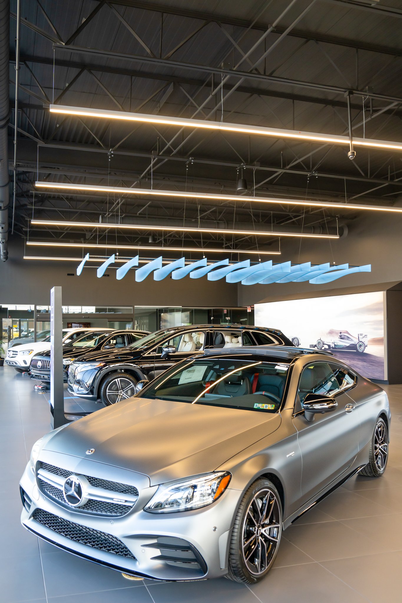 A showroom displaying multiple luxury cars, including a silver Mercedes-Benz convertible in the foreground and various other vehicles in the background, with modern lighting and a large digital screen on the wall.