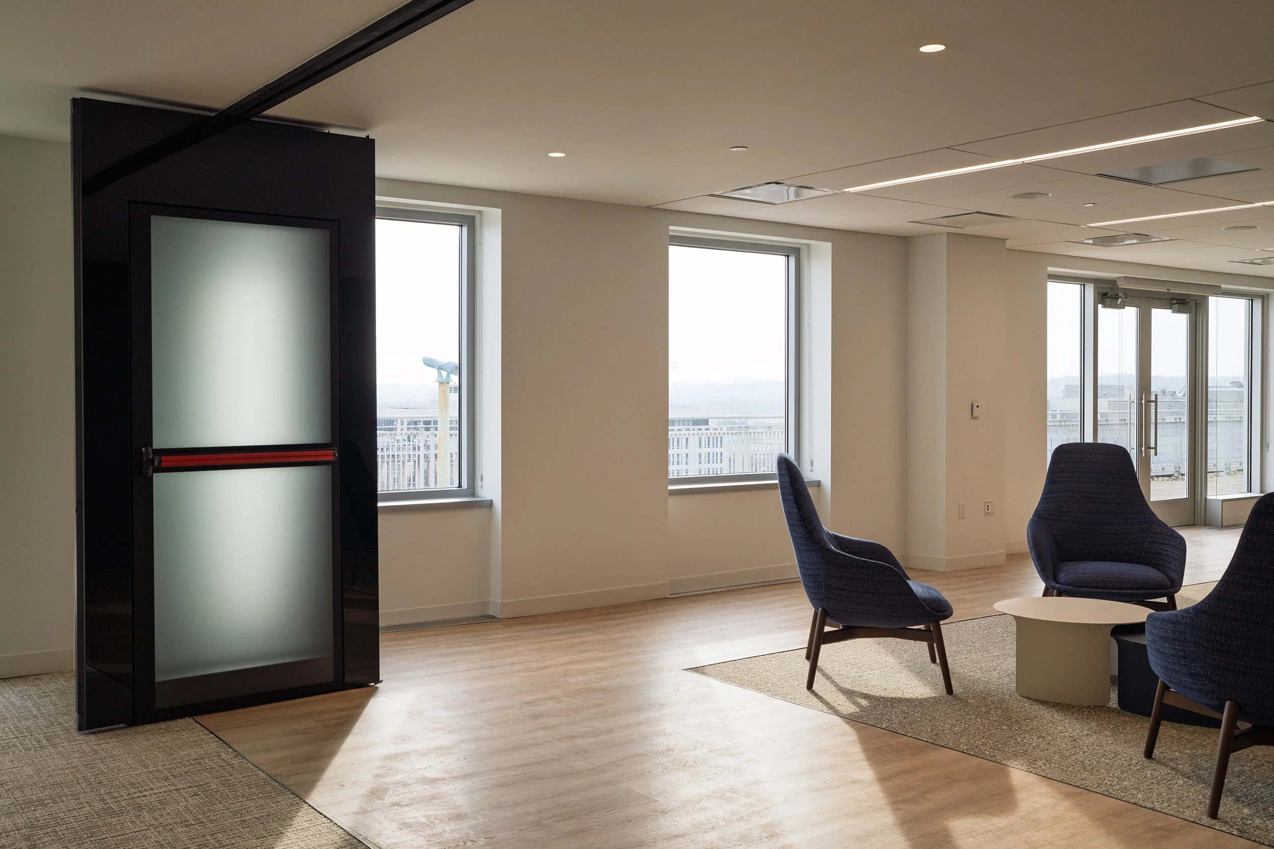 Empty modern lounge area with two dark blue armchairs, a small round white table, large windows, and glass doors leading to an outdoor balcony