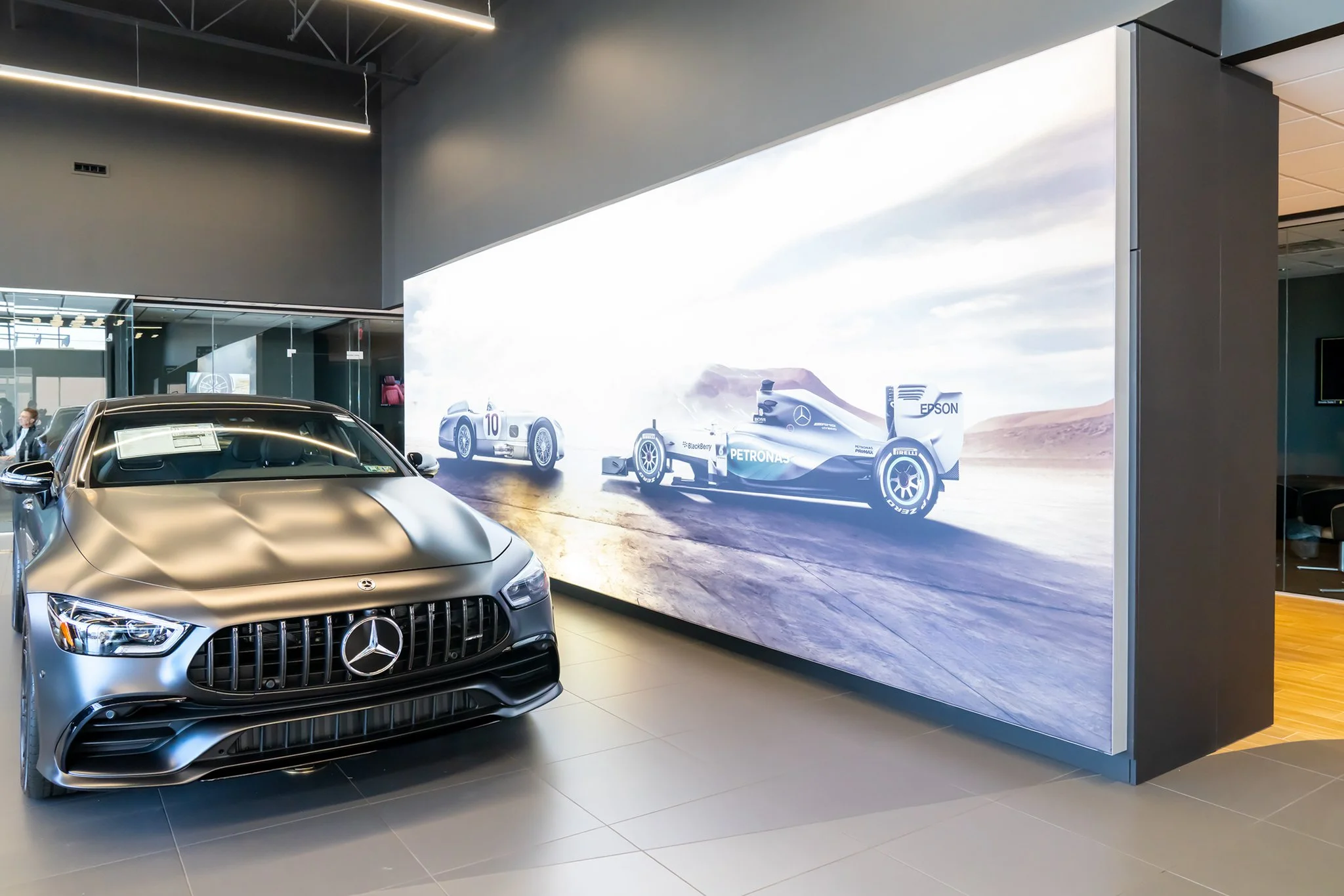 A silver Mercedes-Benz car displayed in front of a large digital screen showing race cars on a desert track.