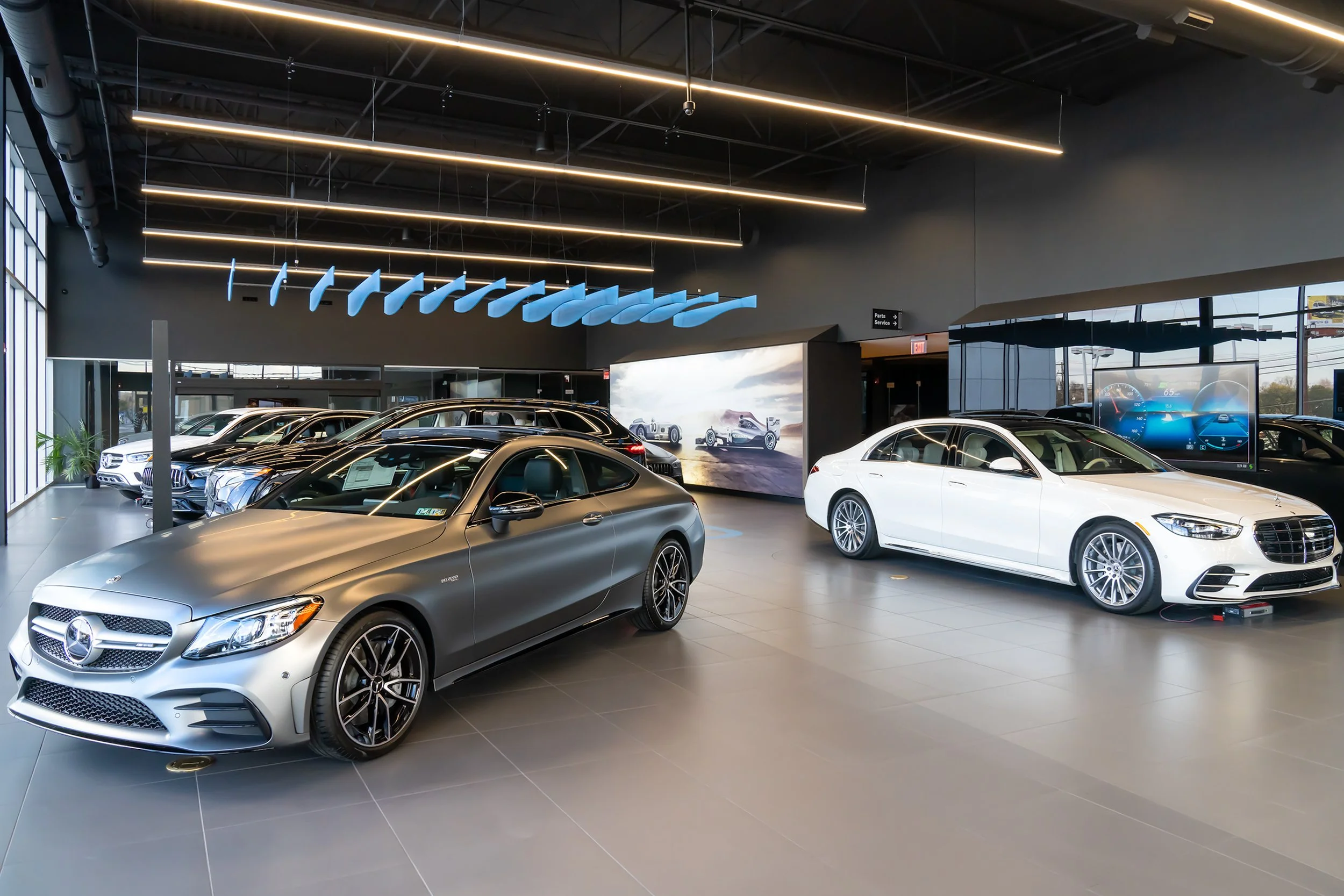 A modern Mercedes-Benz dealership showroom with multiple new luxury cars on display, including a silver coupe and a white sedan, under sleek lighting fixtures with digital screens showing car images and information.