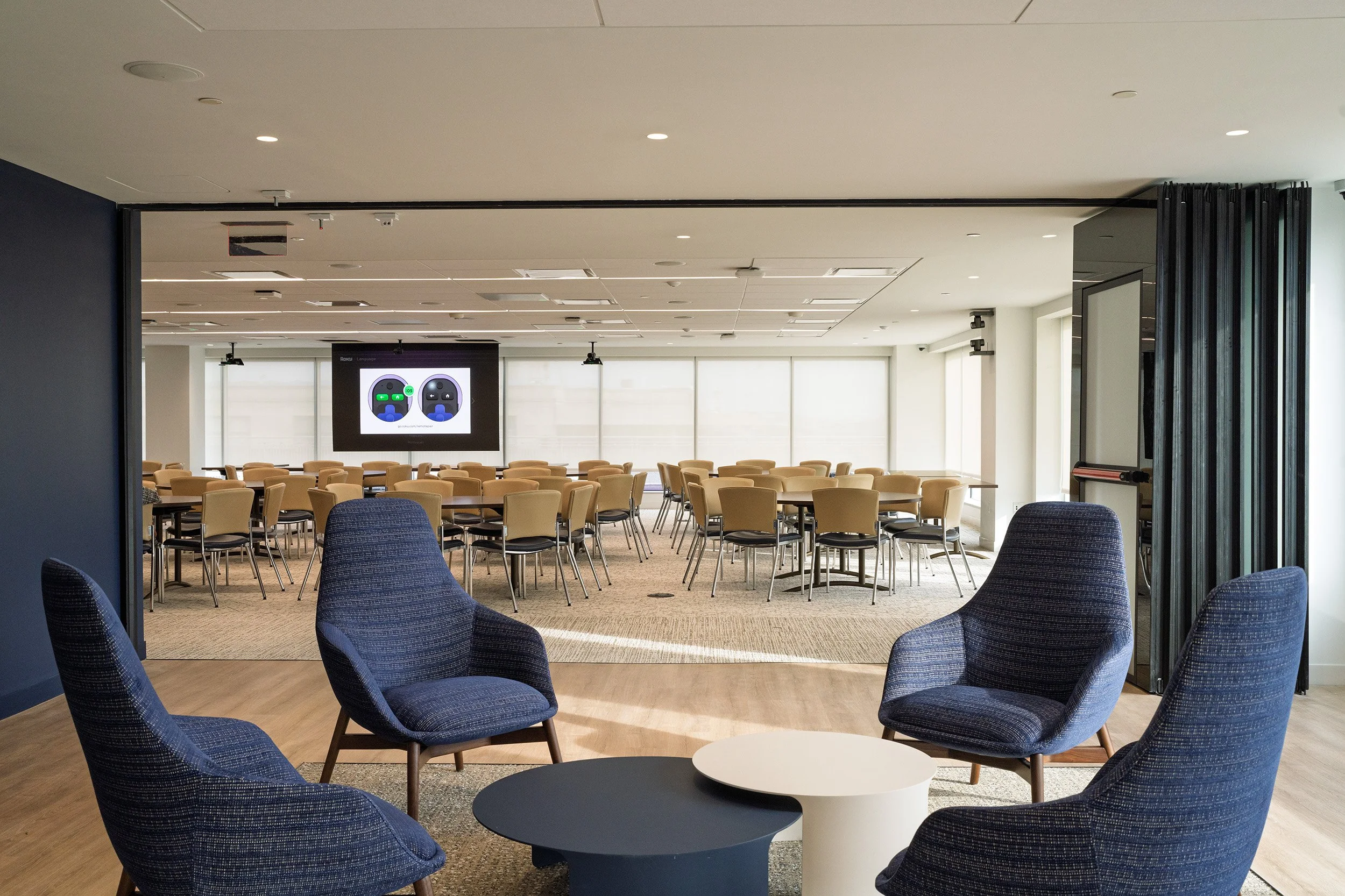 A spacious conference room with a large screen at the front displaying two game controllers, surrounded by multiple tables and beige chairs, and a seating area with four blue upholstered armchairs and a round coffee table in the foreground.