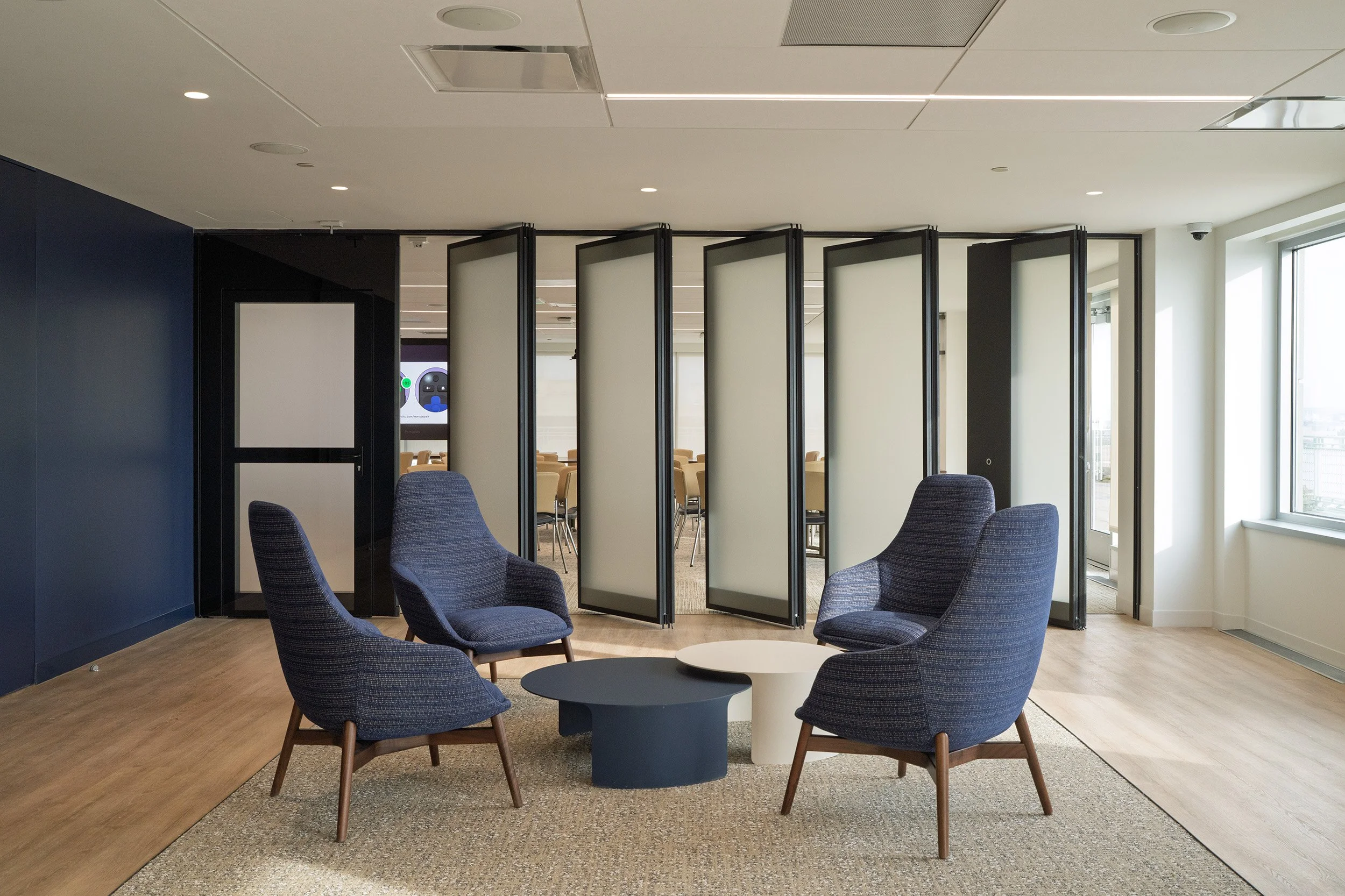 A modern conference room with four blue armchairs arranged around two small round tables, situated on a beige carpet with a larger wooden floor area nearby. In the background, there is a partition with frosted glass panels separating this space from 