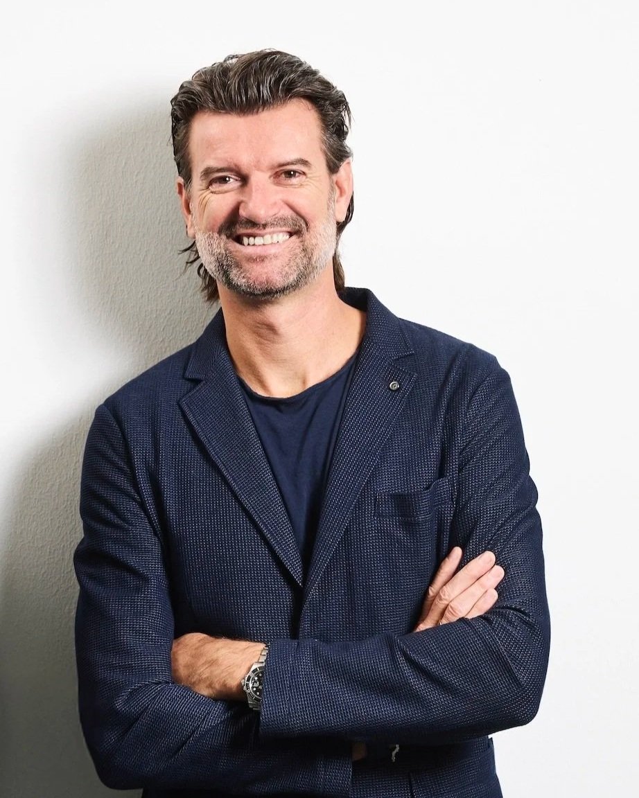 A smiling middle-aged man with gray hair and a beard, wearing a dark blue blazer and a black T-shirt, standing with arms crossed against a plain white wall.