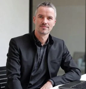 A man with short gray hair and a goatee wearing a black blazer and black shirt, sitting at a desk in an office.