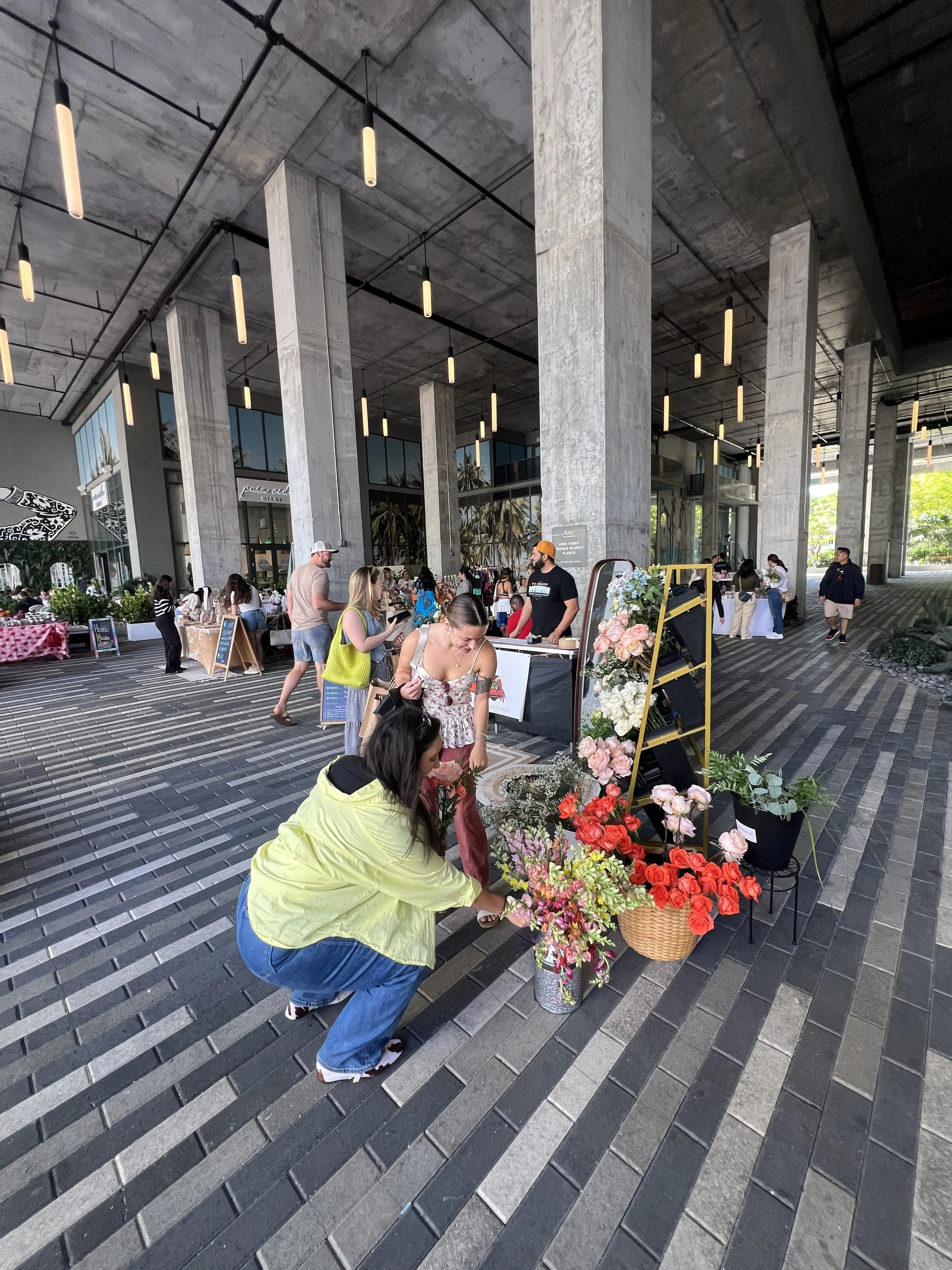 People shopping for flowers at an open-air market under a modern concrete building with hanging lights.