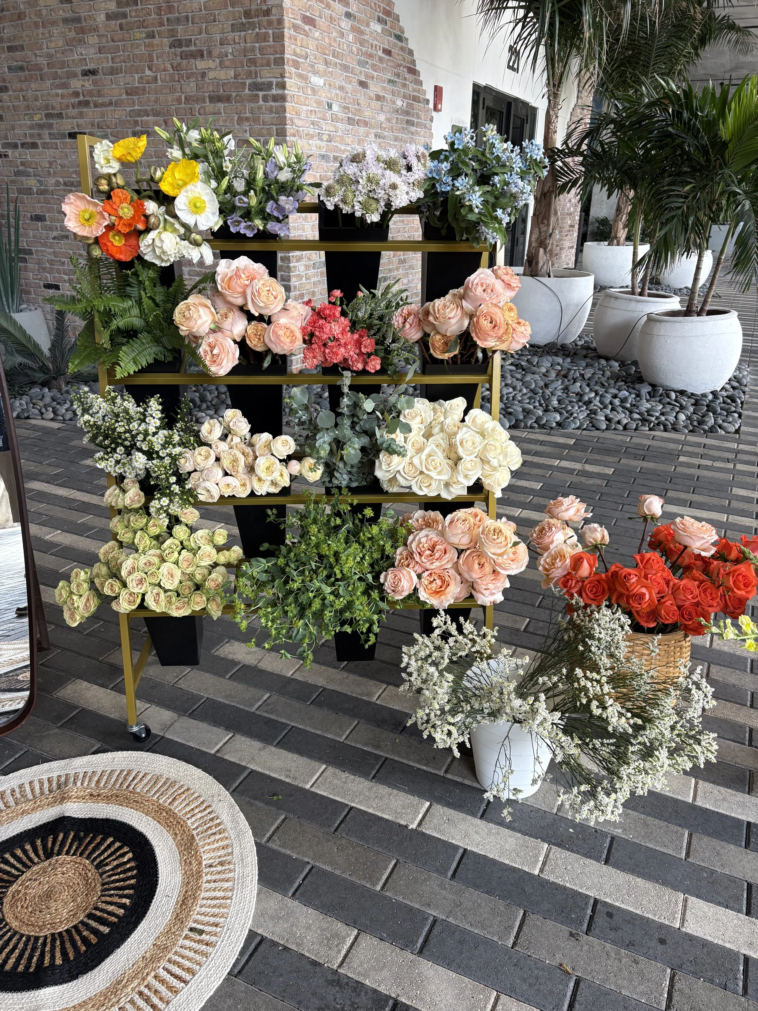 A display of various colorful flowers on a black and gold multi-level stand, with large white pots containing palms and other plants in the background, on a brick-paved outdoor area.