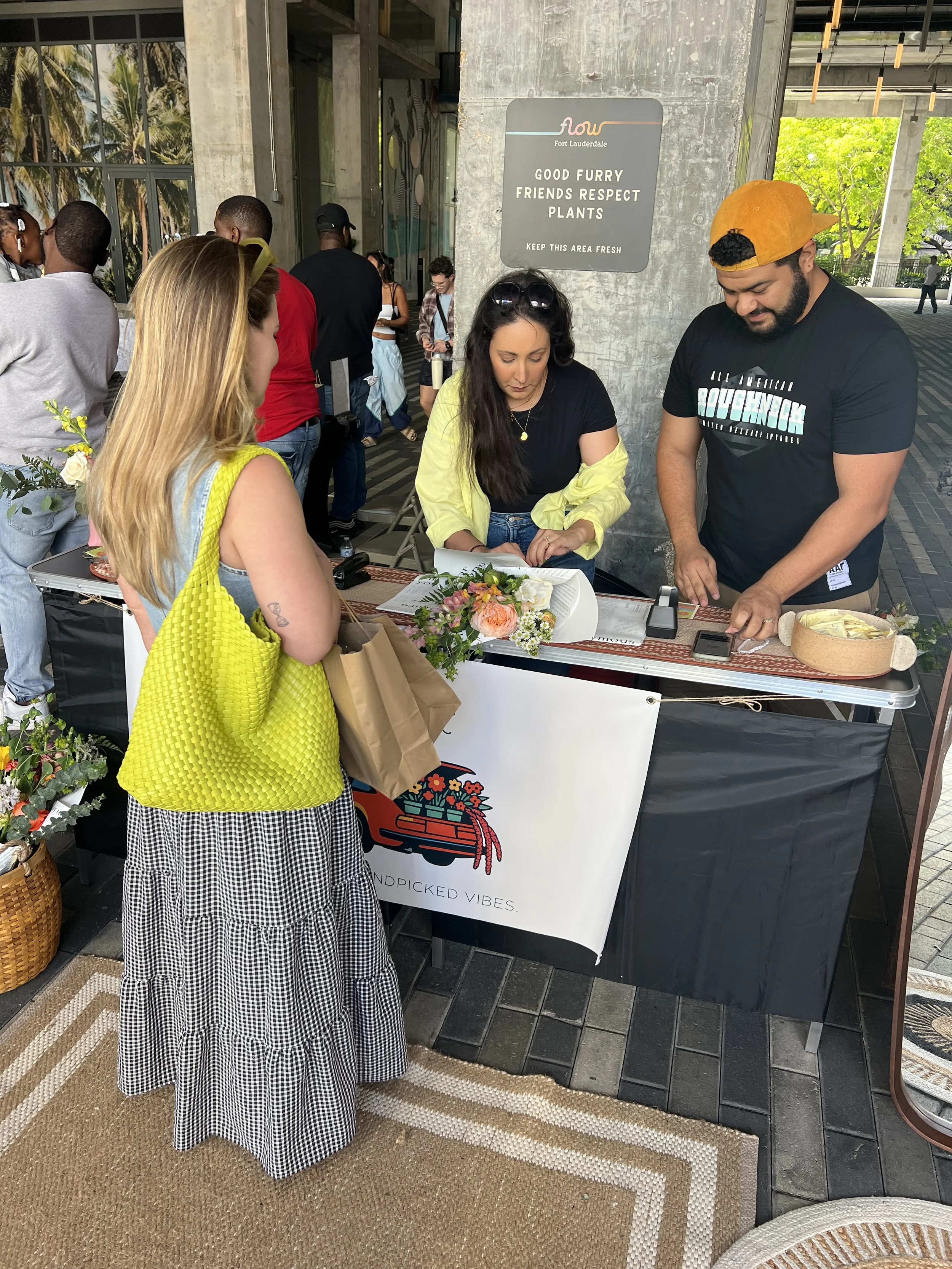 A woman with long blonde hair wearing a yellow handbag and black-and-white checkered skirt is standing in front of a vendor table at an outdoor market, talking to two vendors, a woman with dark hair and a man wearing a yellow cap. The vendors are beh