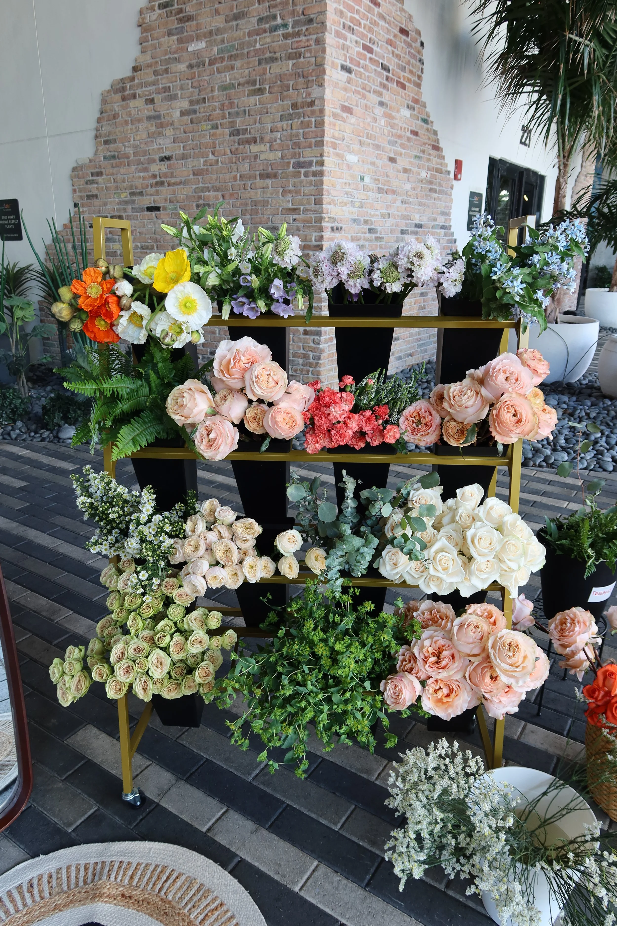 A display of various colorful flowers arranged on black planters and a yellow metal rack outside near a brick wall and potted plants.