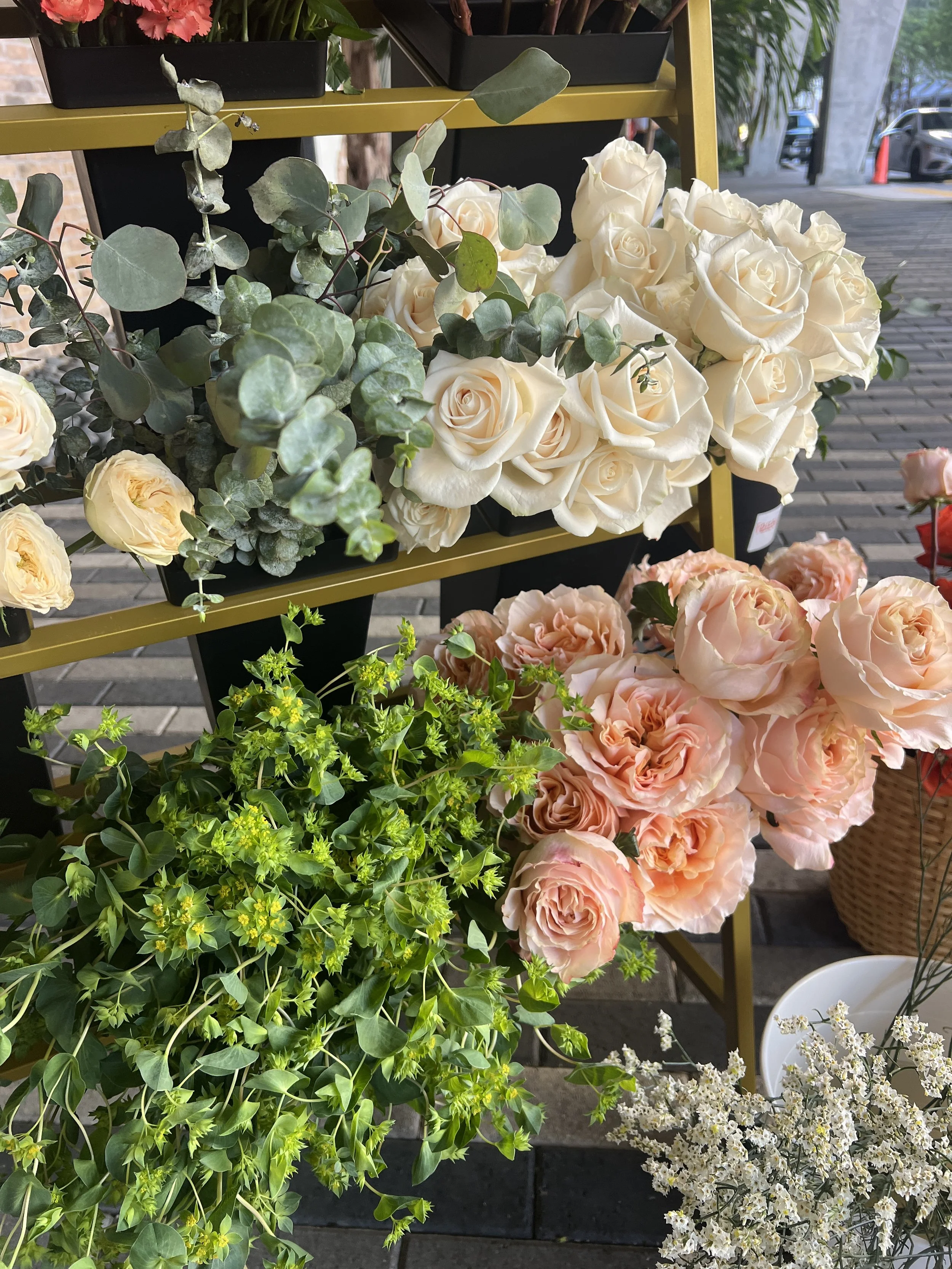 A display of various flowers, including white roses, light pink roses, small white flowers, and green leafy plants, arranged on a metal stand outside.