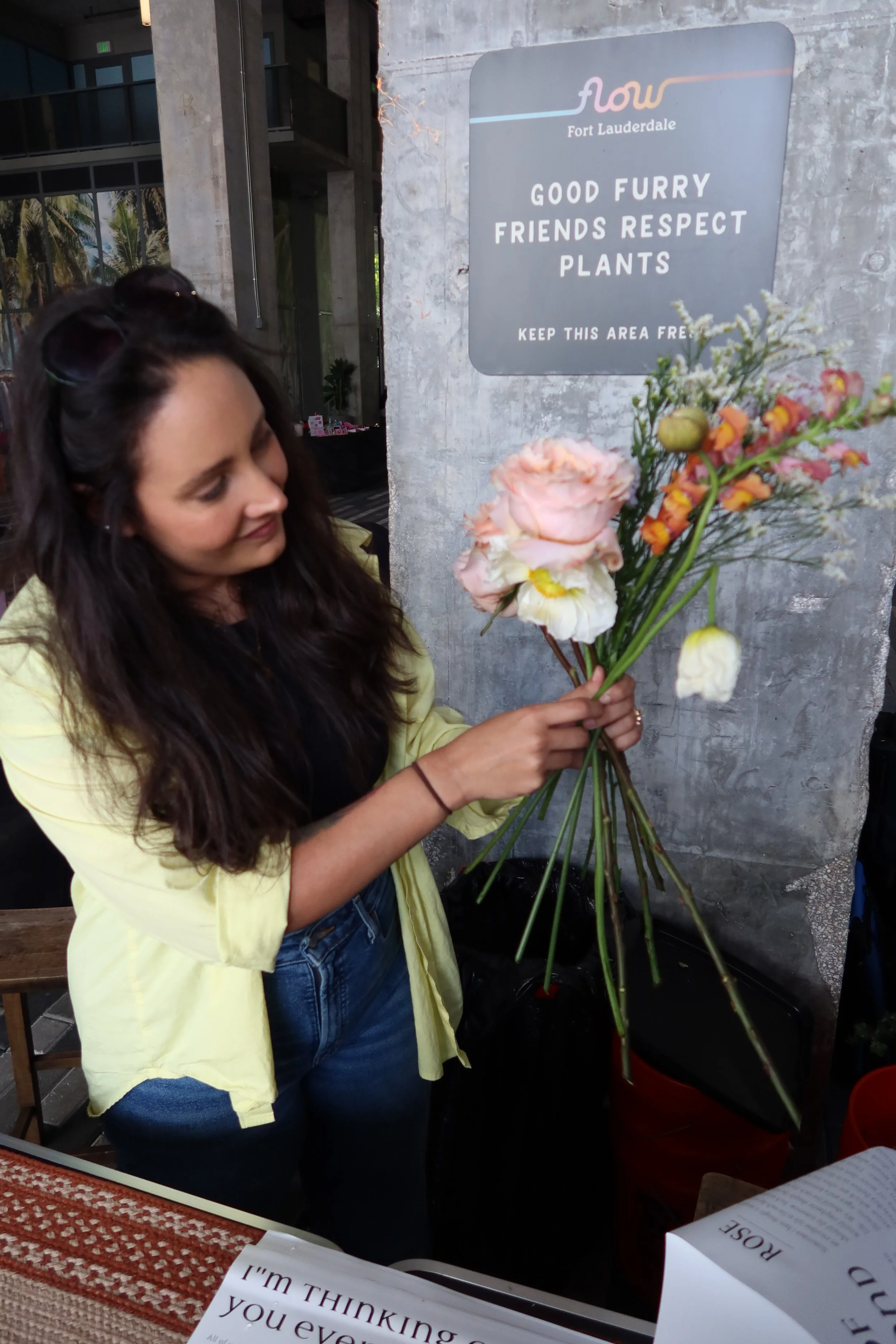 A woman holding a bouquet of mixed flowers, including pink roses, white flowers, and orange blossoms, while standing next to a sign that says 'Good Furry Friends Respect Plants'.