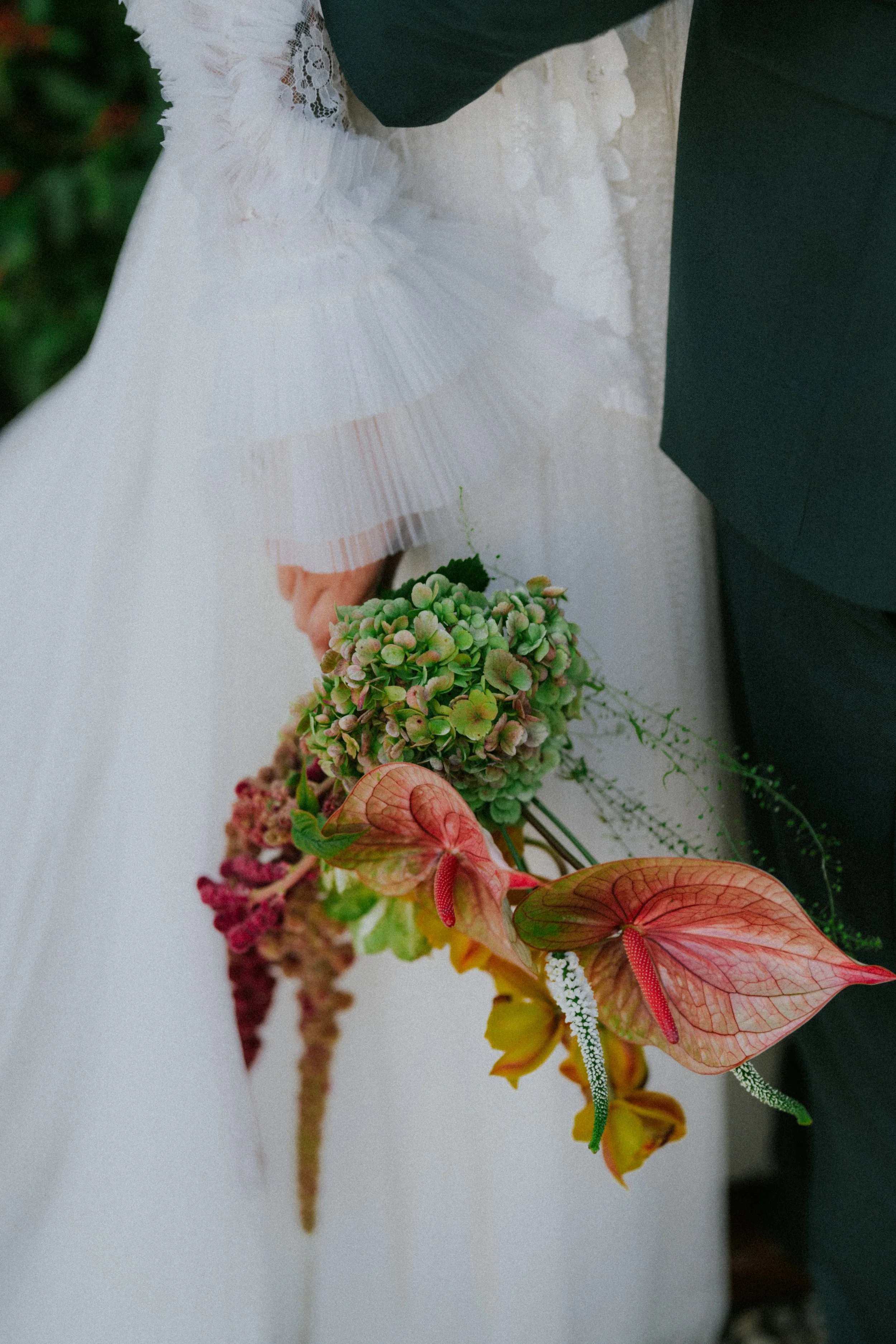 Close-up of a bride and groom holding a wedding bouquet featuring pink anthuriums, green hydrangeas, and other colorful flowers, with the bride wearing a white wedding dress and lace sleeve, and the groom in a dark suit.
