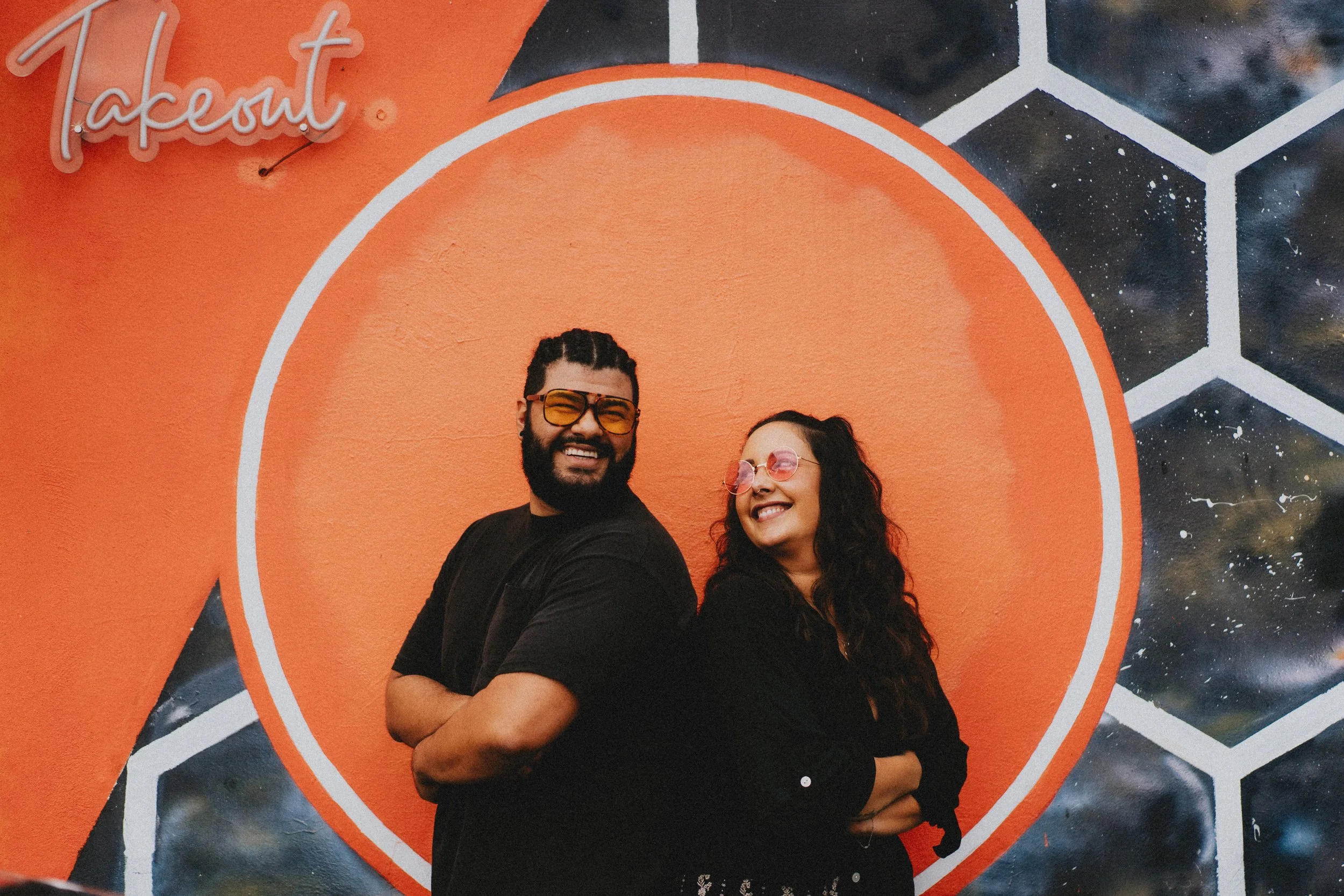 A smiling man and woman stand back to back with arms crossed in front of a colorful mural. The mural features the word 'tapent' in cursive at the top left, a large orange circle, and a hexagon pattern resembling a galaxy or starry sky in black and white.