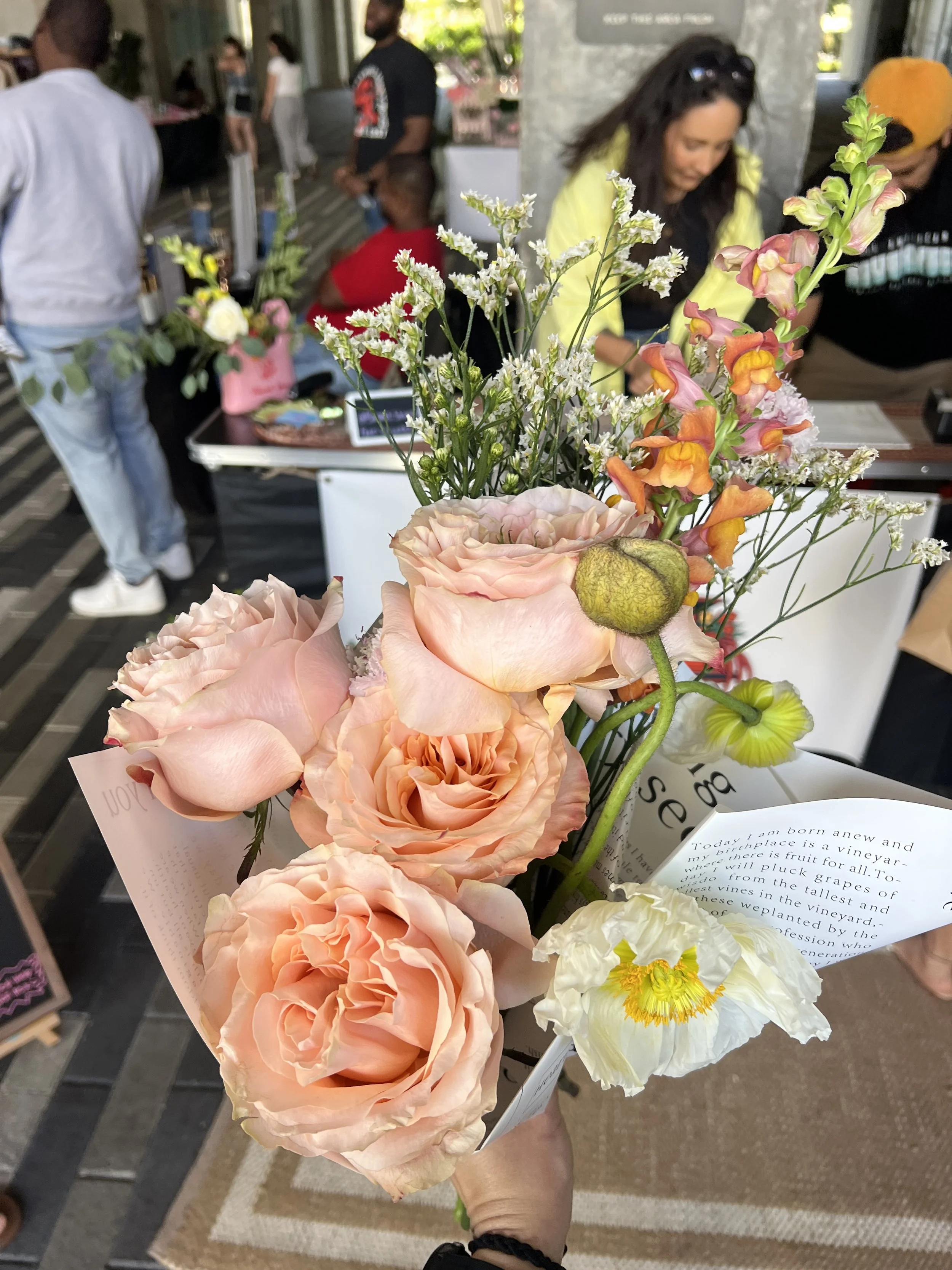Hand holding a bouquet of pink roses, white and yellow poppies, and multicolored snapdragons at a market or event.