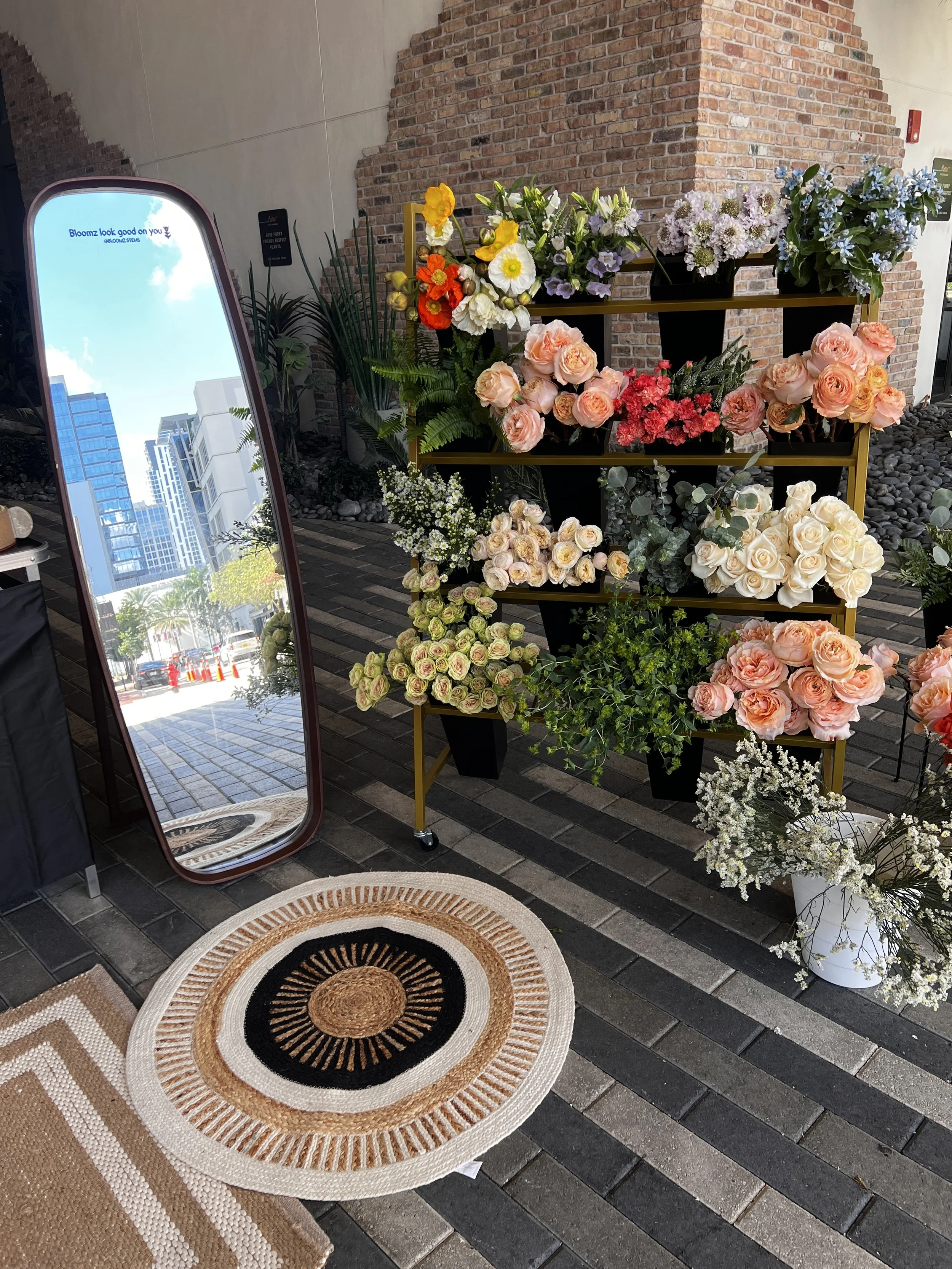 Display of various colorful flowers arranged on a black and gold shelving unit at an outdoor flower shop, with a mirror, a brick wall, and a patterned circular rug on the floor.