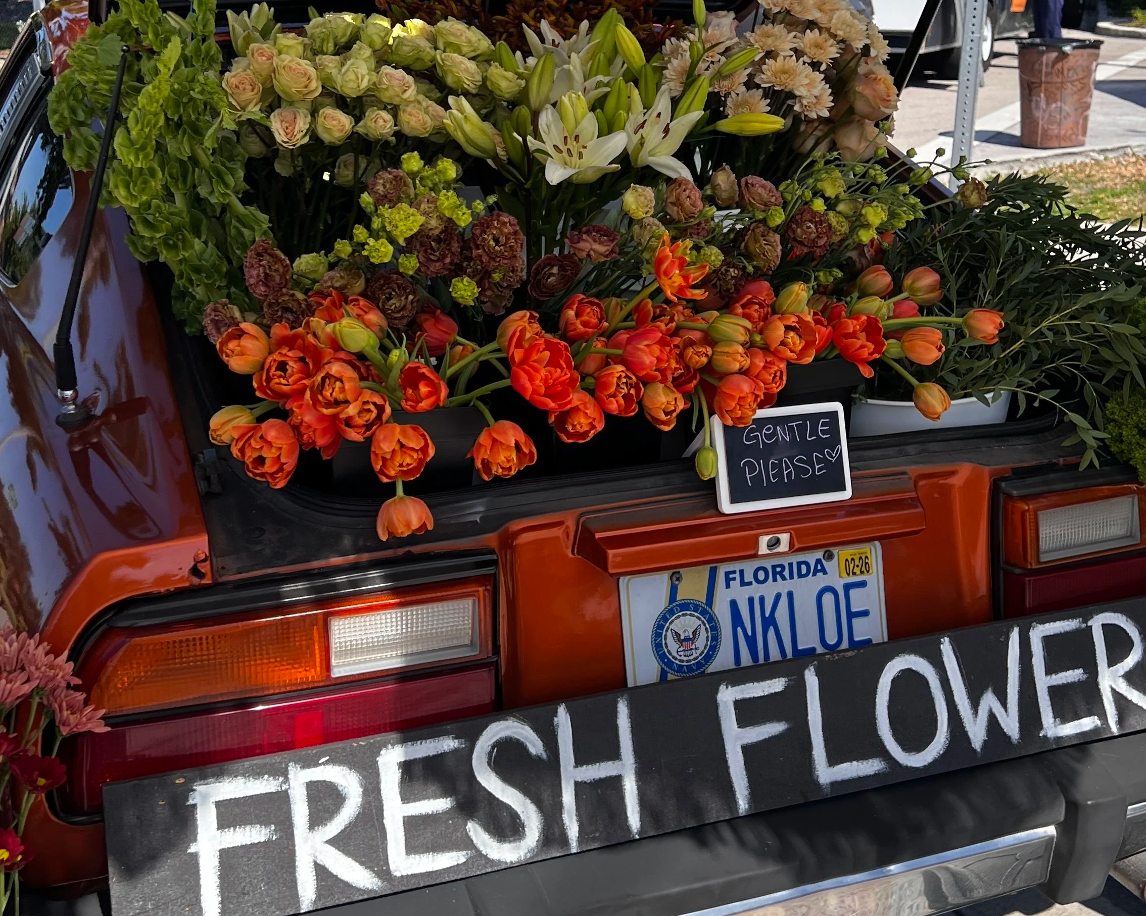 A vintage orange pickup truck filled with various fresh flowers in the truck bed, including orange tulips, white lilies, and green hydrangeas. A small chalkboard sign saying 'GENTLE PLEASE' is attached to the truck, and a large black sign with white 