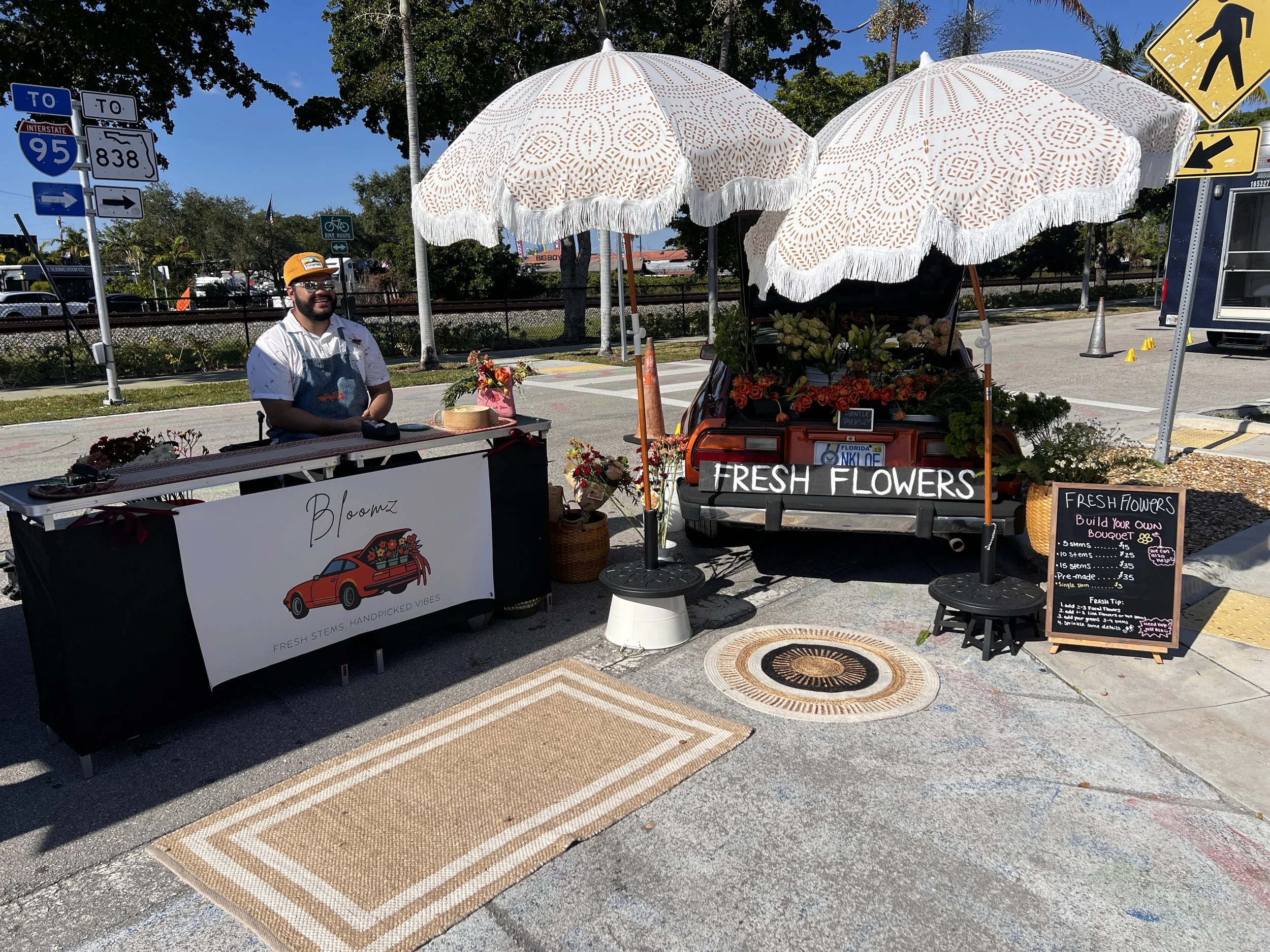 A flower stand named 'Bloomz' is set up on a sidewalk with a banner displaying a red car carrying flowers. The stand has a white umbrella, with an additional car showcasing fresh flowers, and a chalkboard sign listing flower pricing. An employee wear