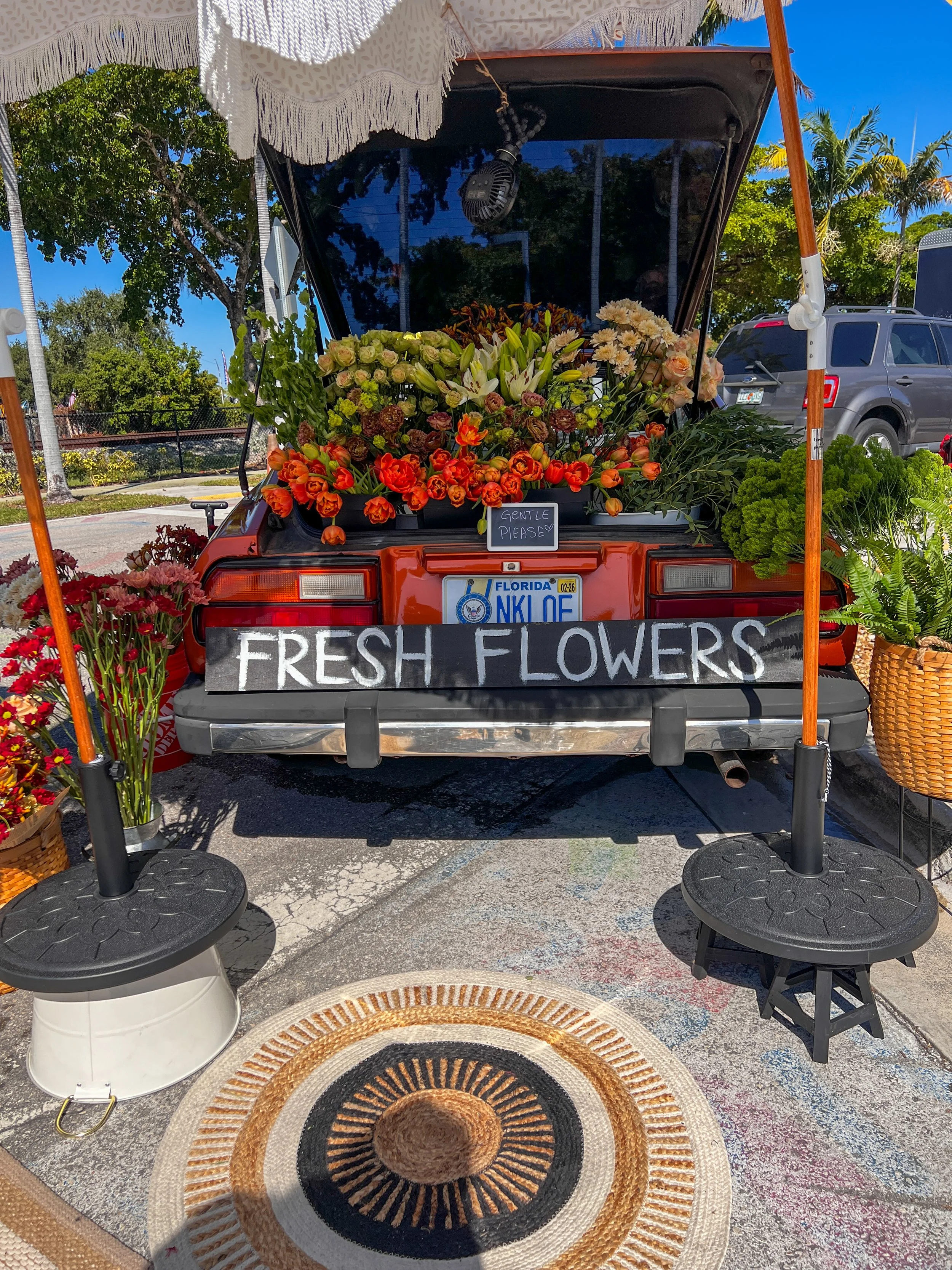 A flower vendor stand with a sign reading 'Fresh Flowers' and a variety of colorful fresh flowers for sale, set up on the back of an orange vehicle outside on a sunny day.