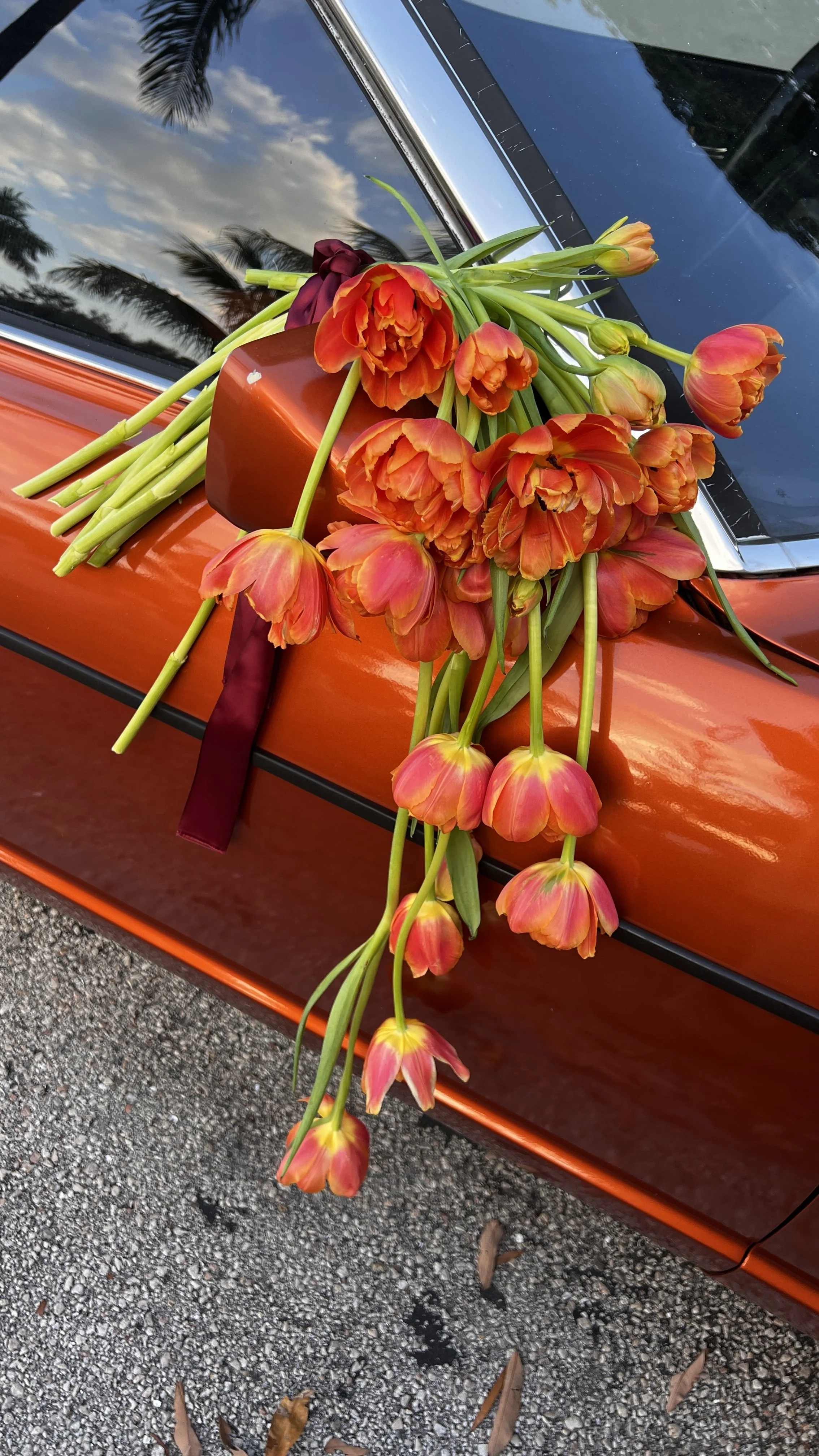 Orange tulips arranged on the hood of a shiny orange car, with a small brown vase and a maroon ribbon in the decoration, reflecting partly cloudy sky and palm trees in the background.