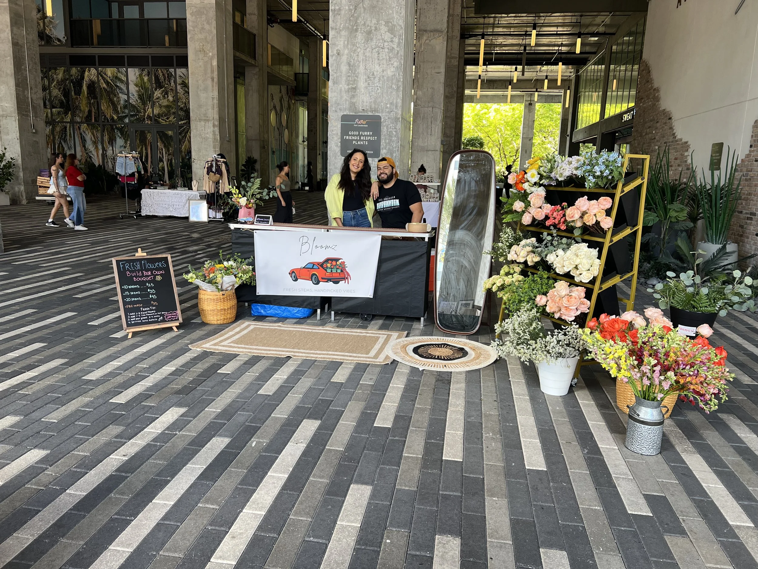 A flower stand with various bouquets and a sign that reads build your own bouquet, set up in a spacious indoor area with concrete walls, large window, and people walking in the background. Two people stand behind the stand smiling at the camera.