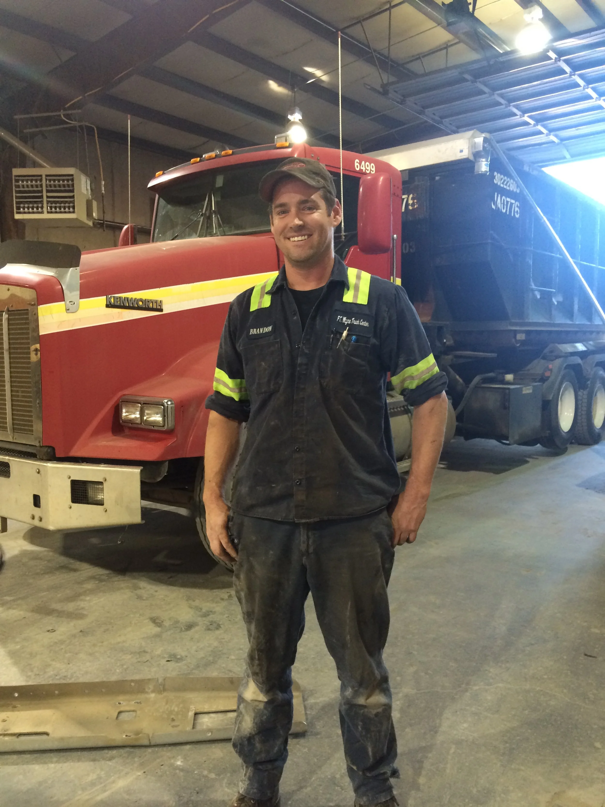 A man in work clothes, with dirty hands and pants, standing inside a fire station garage. He is smiling and wearing a dark shirt with reflective strips, a cap, and has pens in his chest pocket. Behind him is a large red fire truck and a blue trailer.