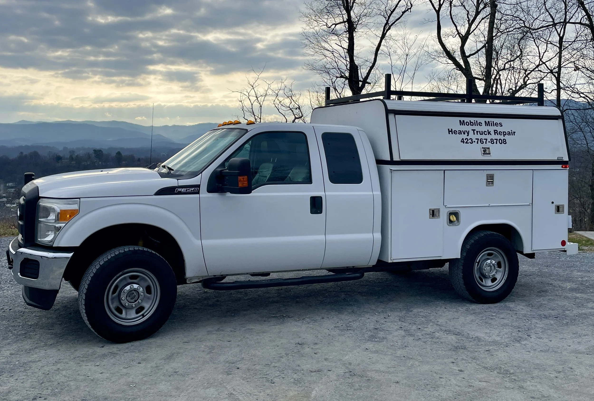 White utility truck with equipment compartments parked on gravel with mountains and cloudy sky in the background.