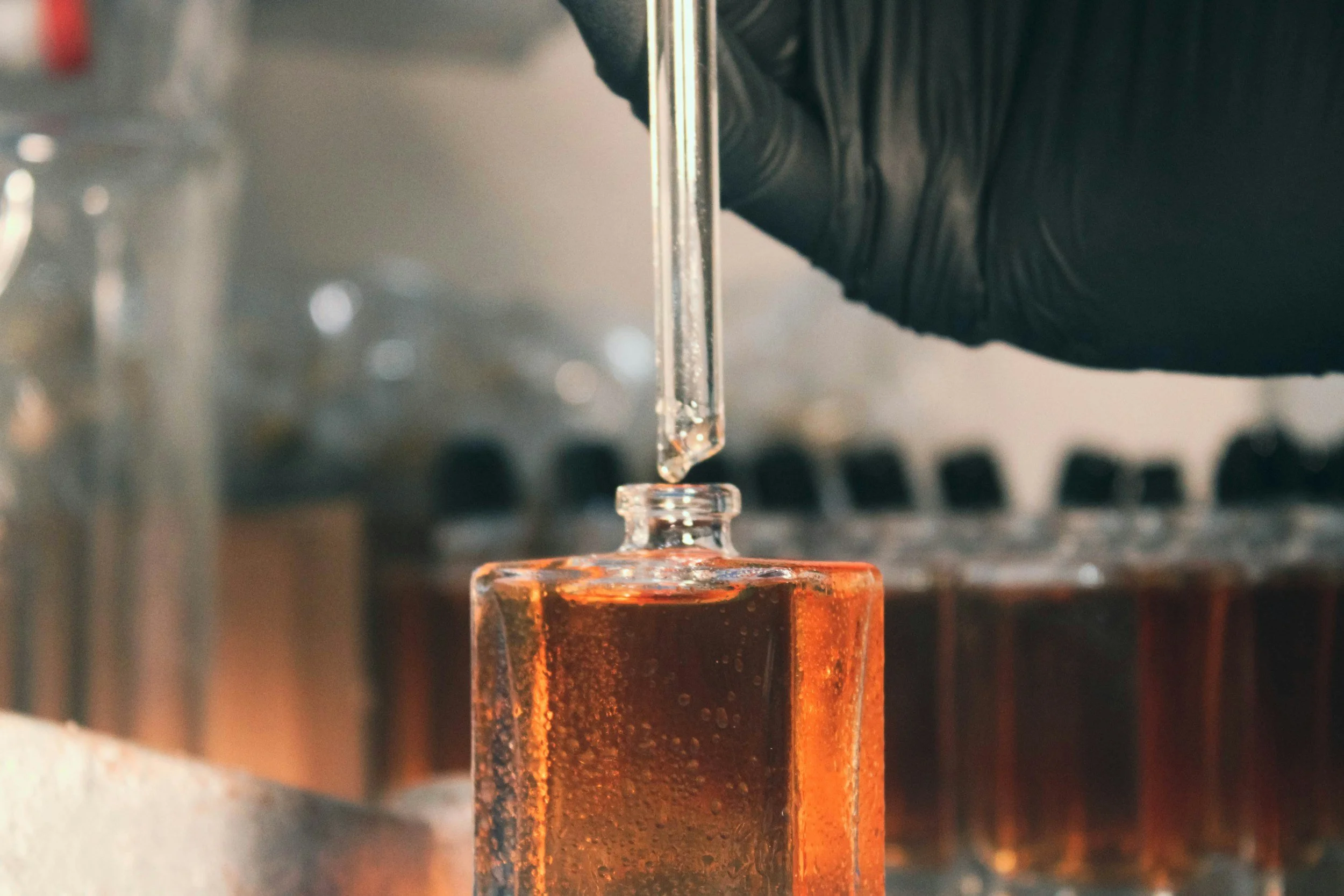 A person wearing black gloves adding a liquid drop into a small glass bottle with amber-colored liquid, in a laboratory setting.
