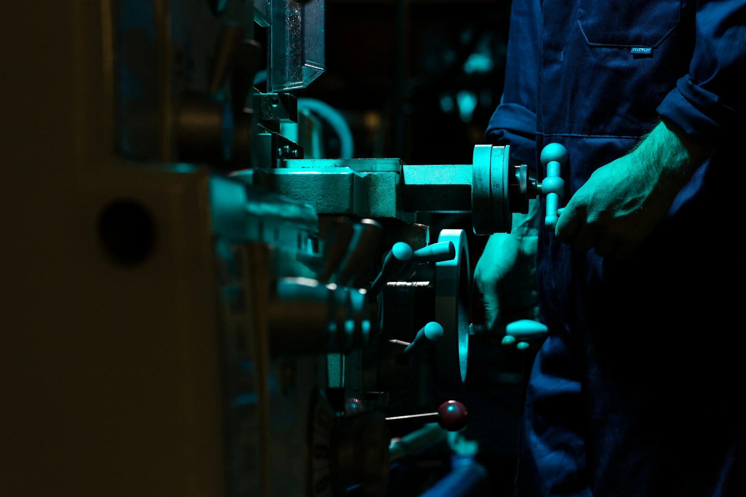 A person wearing blue gloves operating a metal machine in a dimly lit workshop.