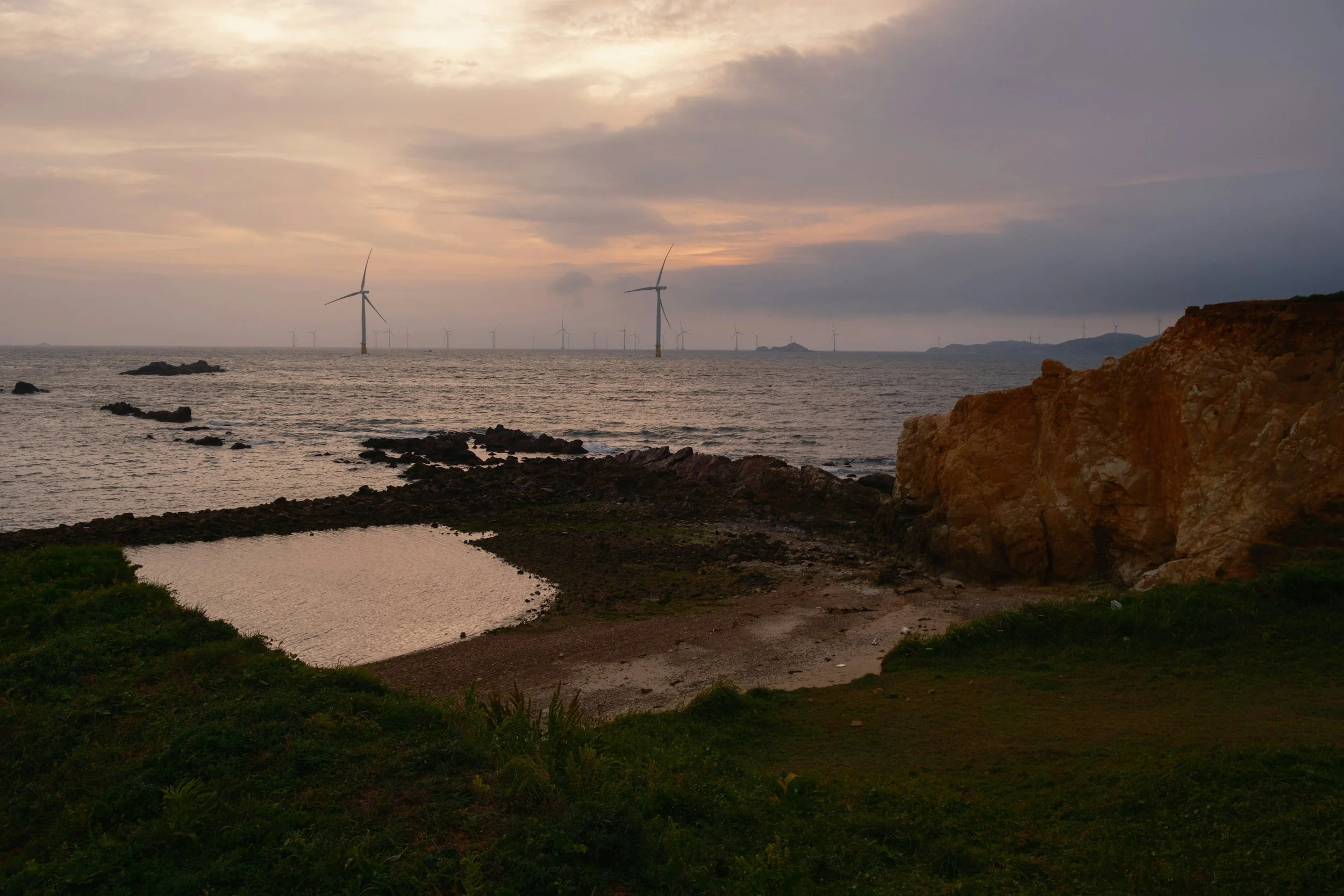 A coastal scene at sunset with wind turbines in the distance, rocky shoreline, a small shallow pool of water, grassy area, and a large rocky cliff.