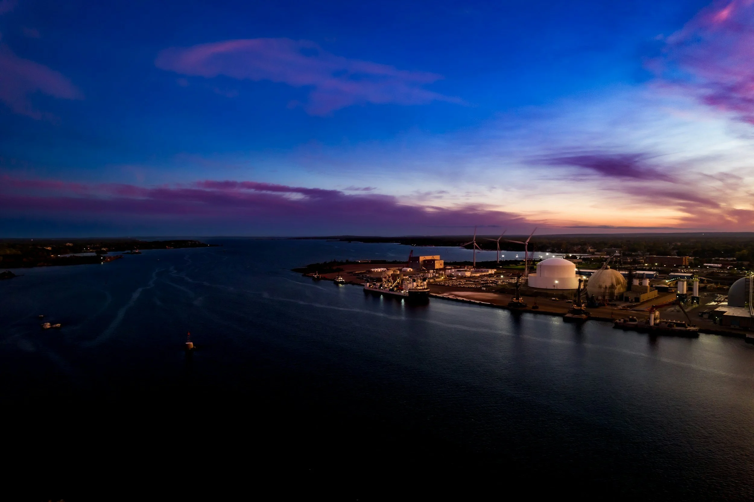 Aerial view of a harbor at sunset with wind turbines, storage tanks, and a body of water.