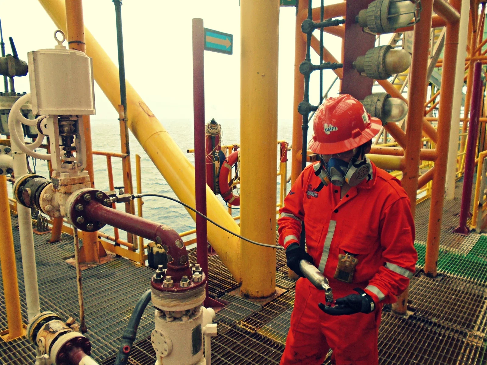 Offshore worker in orange safety suit and red helmet working on equipment on an oil platform at sea.