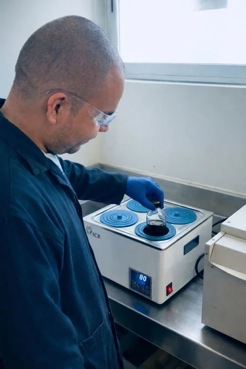 A scientist wearing safety glasses and gloves adding a liquid to a petri dish on a hot plate in a laboratory.