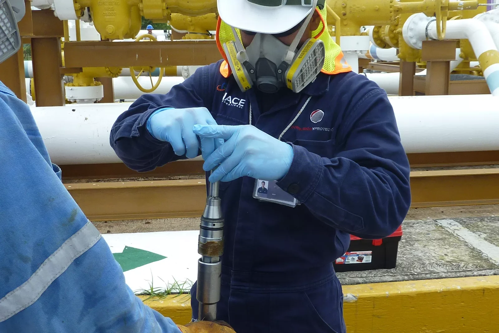 Worker wearing protective goggles, mask, gloves, and a blue jumpsuit, handling a piece of equipment in an industrial setting with pipes and machinery in the background.