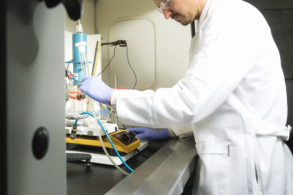 A scientist in a white lab coat and purple gloves working with laboratory glassware and equipment in a lab setting.