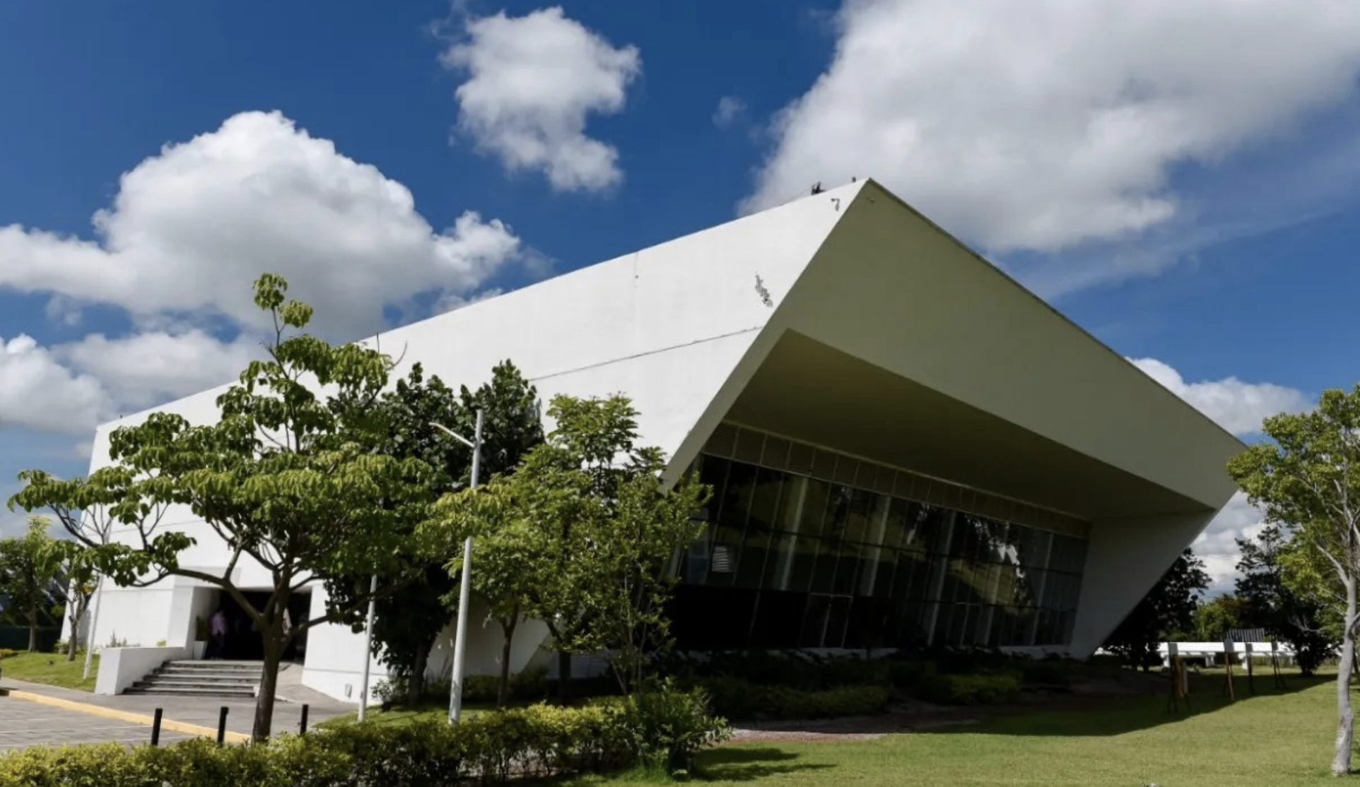 Modern white building with an angled roof, surrounded by green trees and grass under a blue sky with clouds.