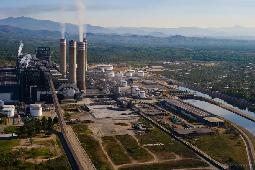 An industrial power plant with multiple smokestacks emitting smoke, surrounded by mountains and greenery.