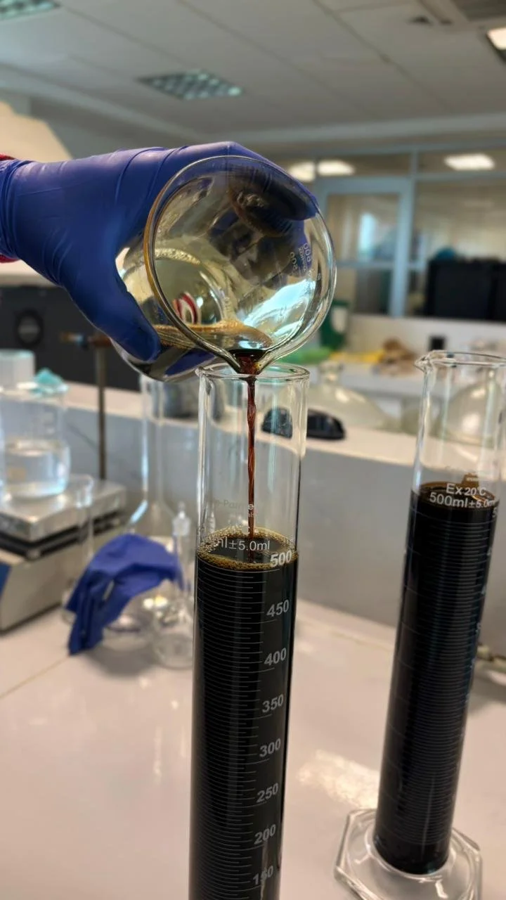 A scientist is pouring a dark liquid from a small beaker into a graduated cylinder in a laboratory setting.