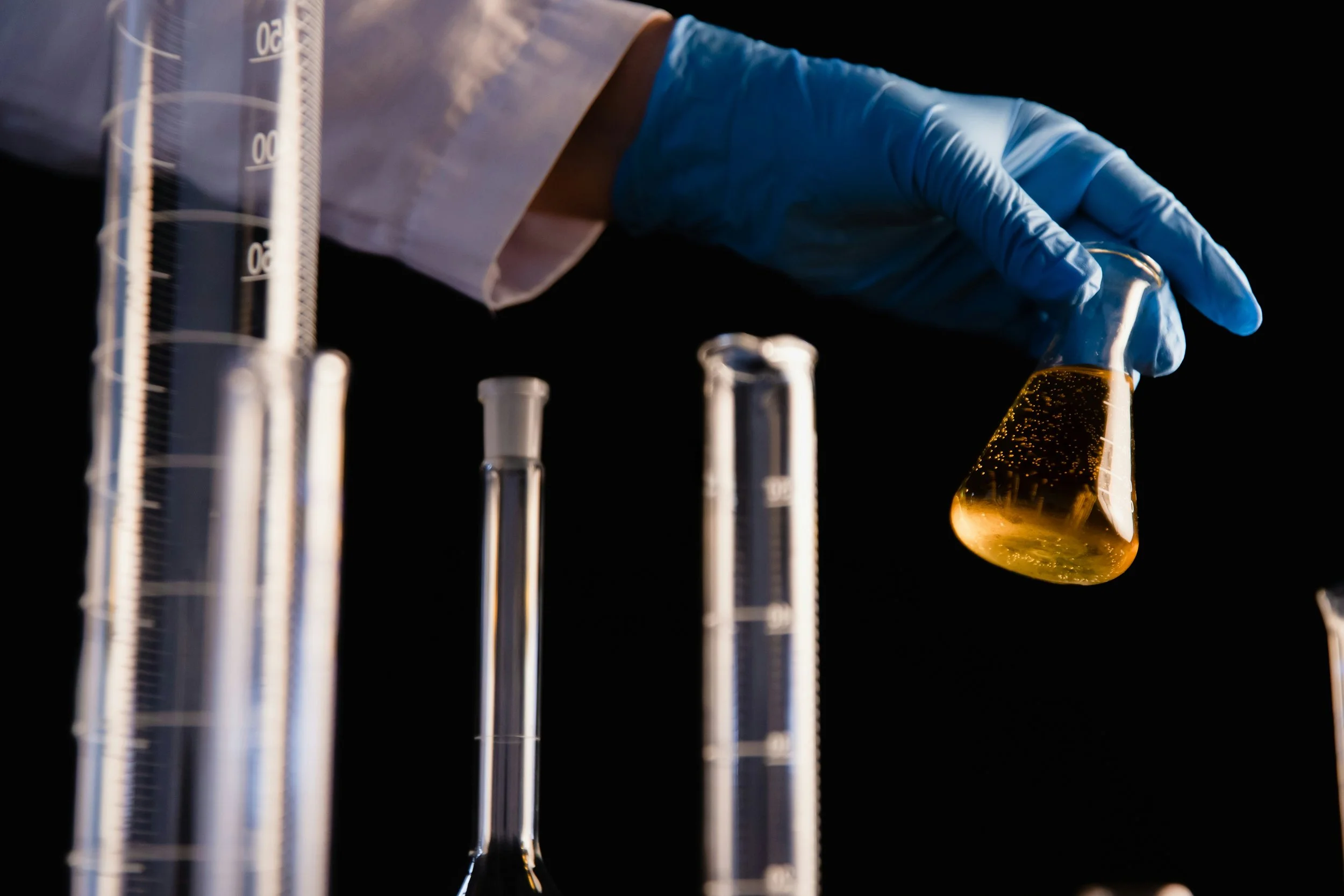 A scientist wearing a blue glove holding a conical flask with yellowish liquid, with several graduated cylinders placed on a black background.