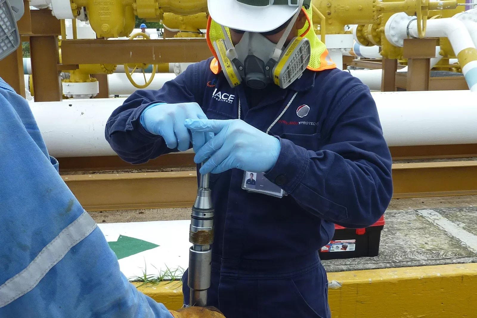 A worker wearing a respirator mask, gloves, and a safety vest is assembling or inspecting a piece of industrial equipment in an outdoor setting with yellow pipes and machinery in the background.