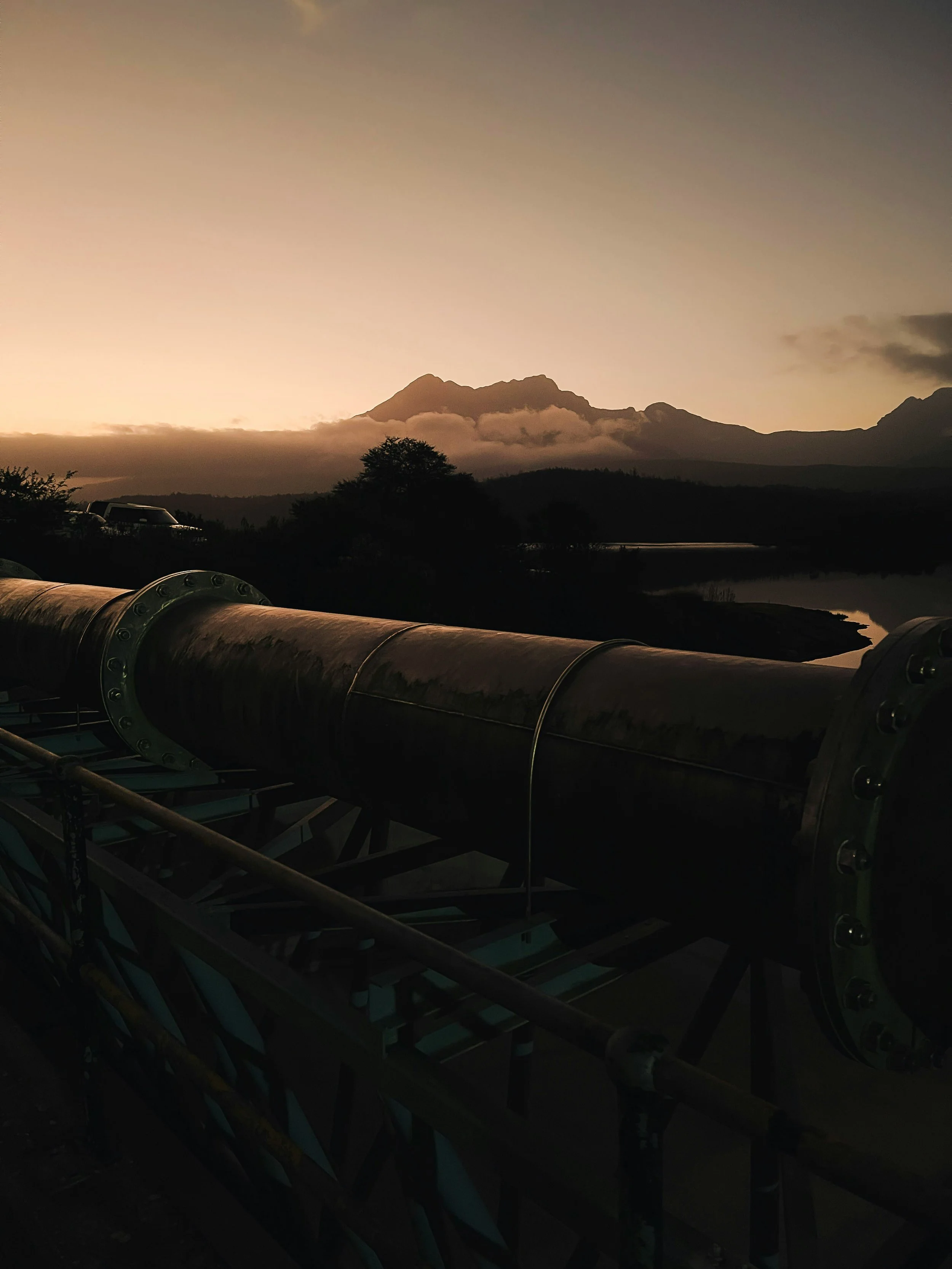 A large industrial pipe on a support structure with a mountain and sunset in the background.