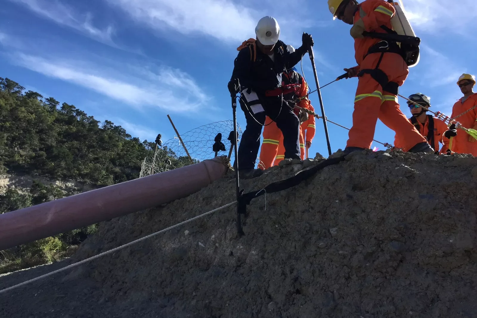 Firefighters in orange uniforms and helmets secure a fallen utility pole and power lines on a dirt hill, with safety fencing, under a blue sky with clouds.