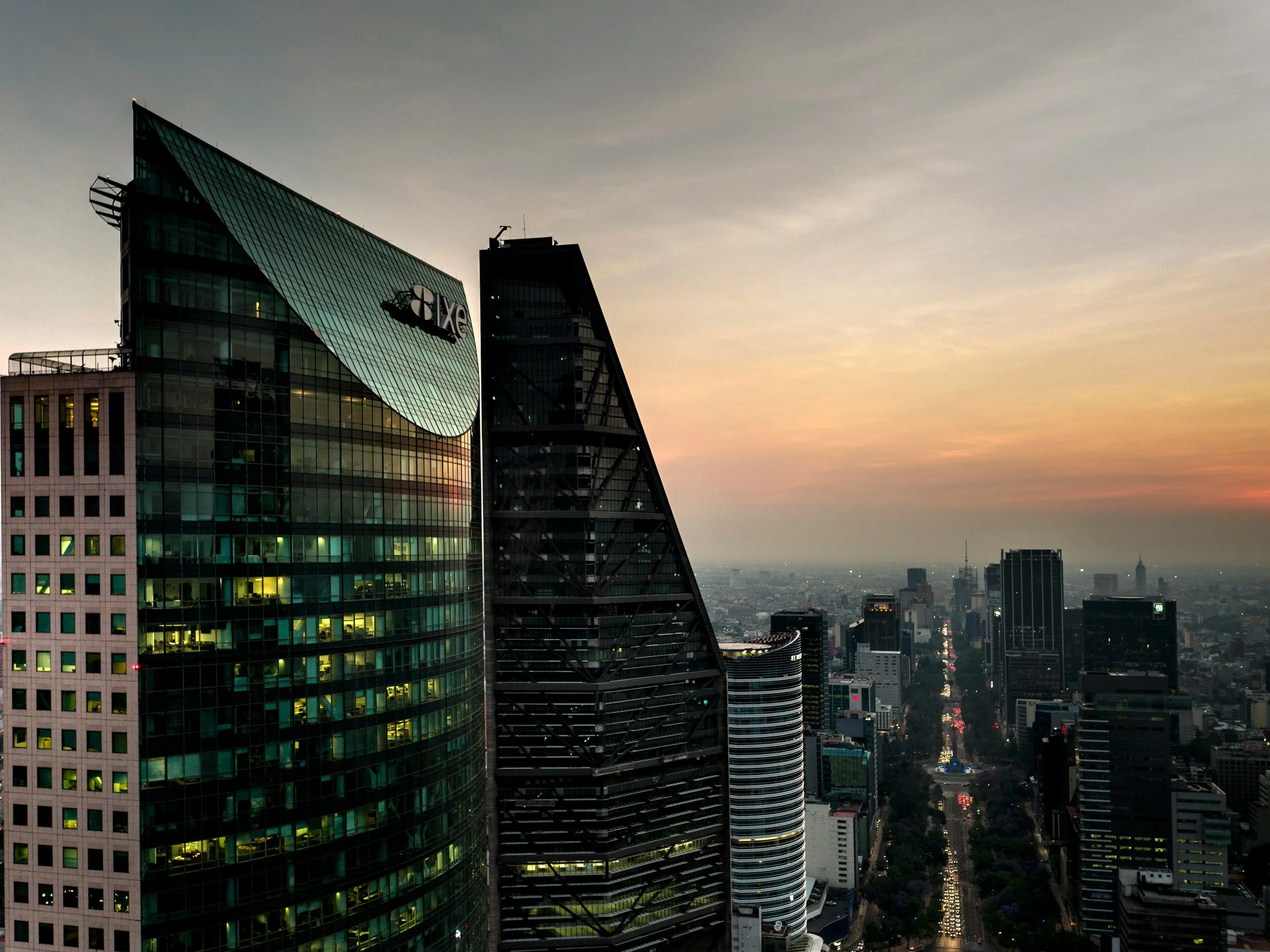 City skyline at sunset showing tall modern skyscrapers, including a glass building with the 'AXE' logo, and a street with moving lights extending into the distance.